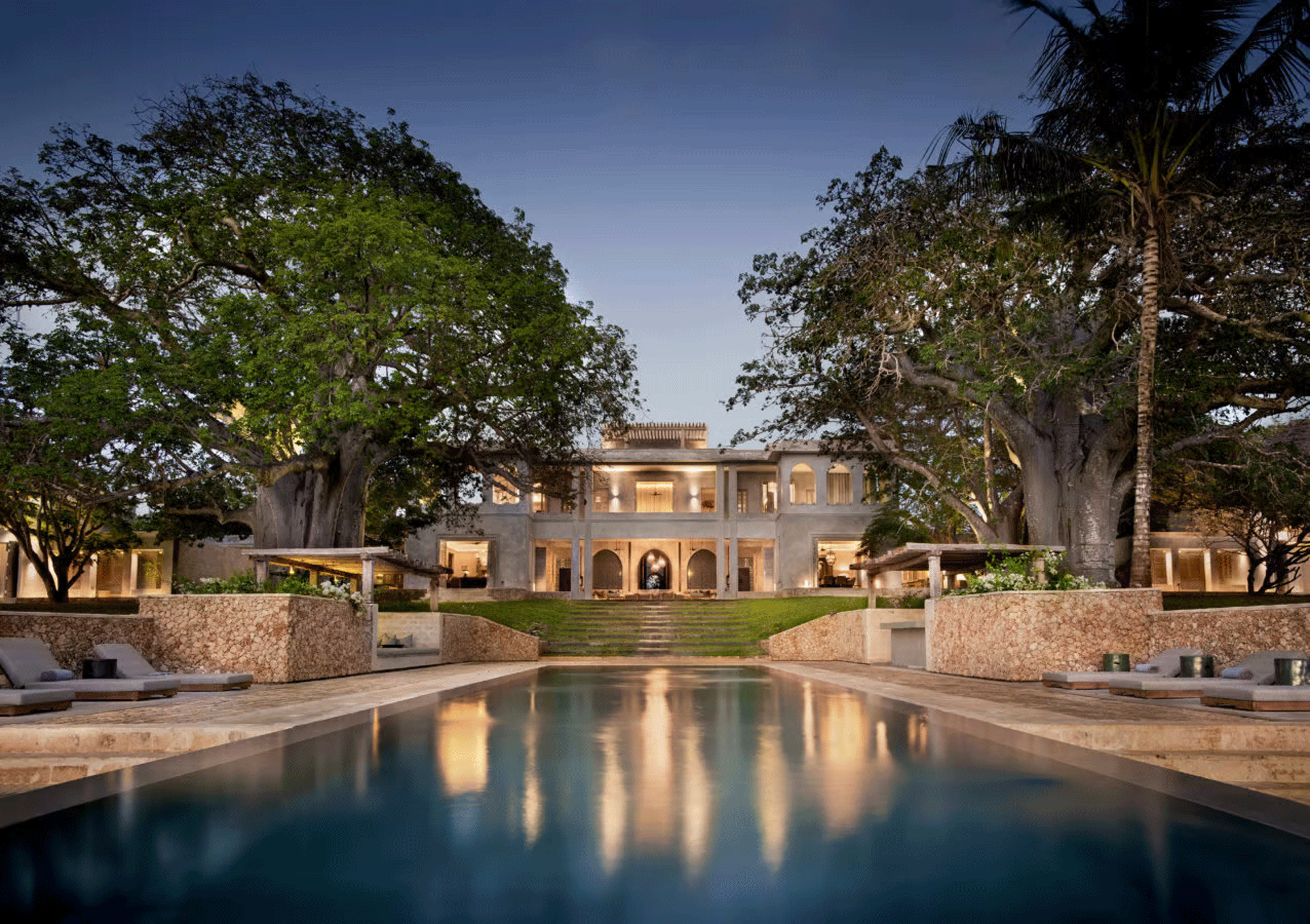 Dusk view of a low villa mirrored in a still pool beneath large spreading trees.