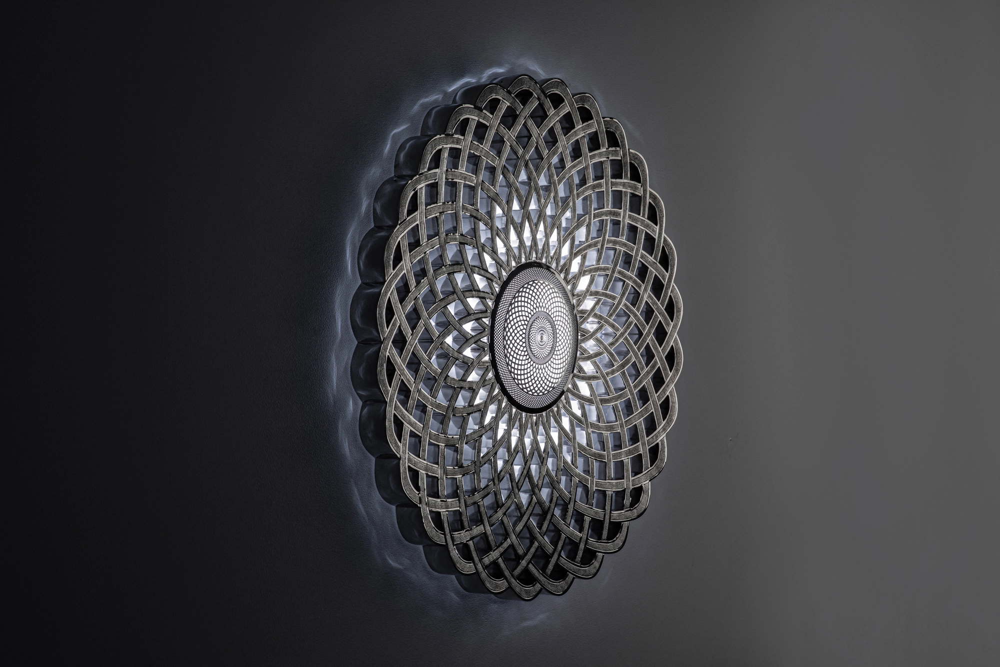 A circular openwork metal disc with fine concentric lattice, mounted on a grey wall in dim light, Infinity 818.
