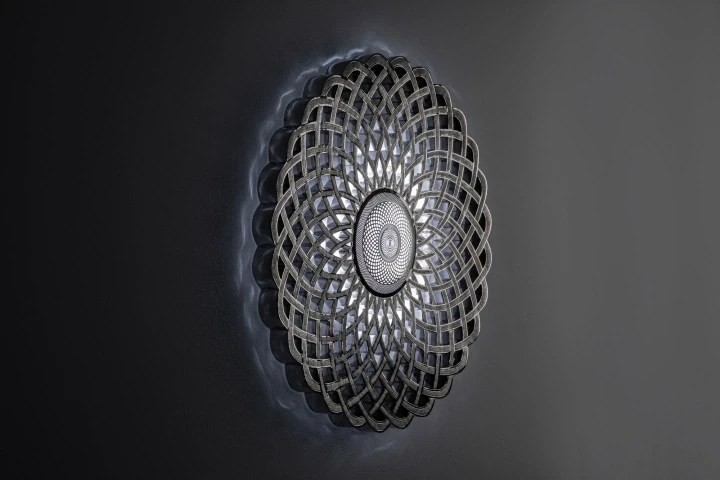 A circular openwork metal disc with fine concentric lattice, mounted on a grey wall in dim light, Infinity 818.