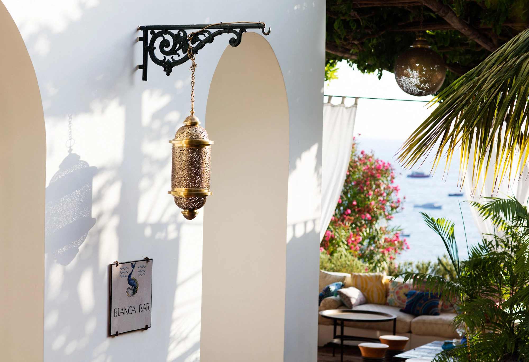 White archway with a hanging gold lantern, a small sign and a palm set against the sea.