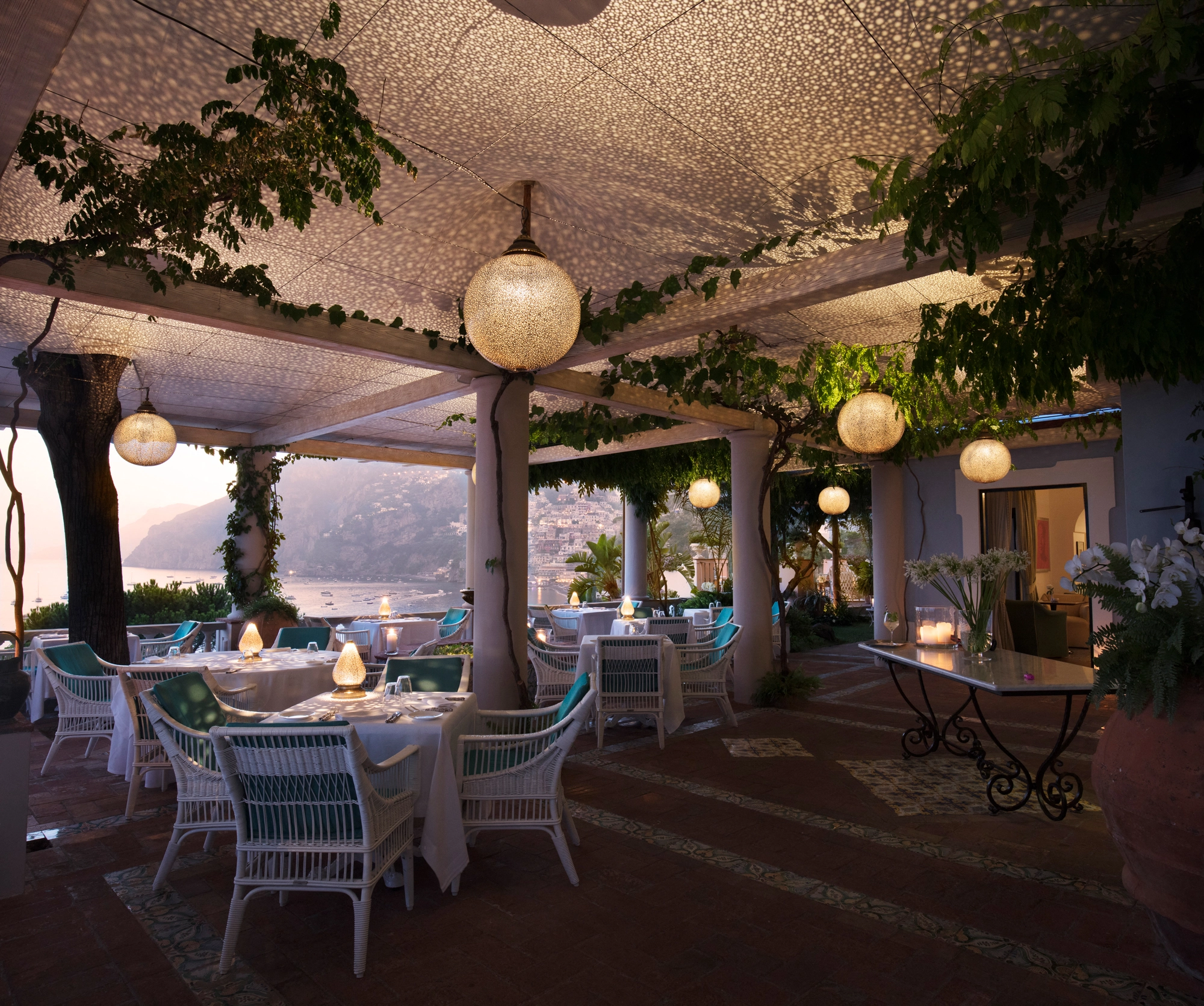 Evening terrace dining beneath a pierced ceiling with a globe lantern and a coastal view.