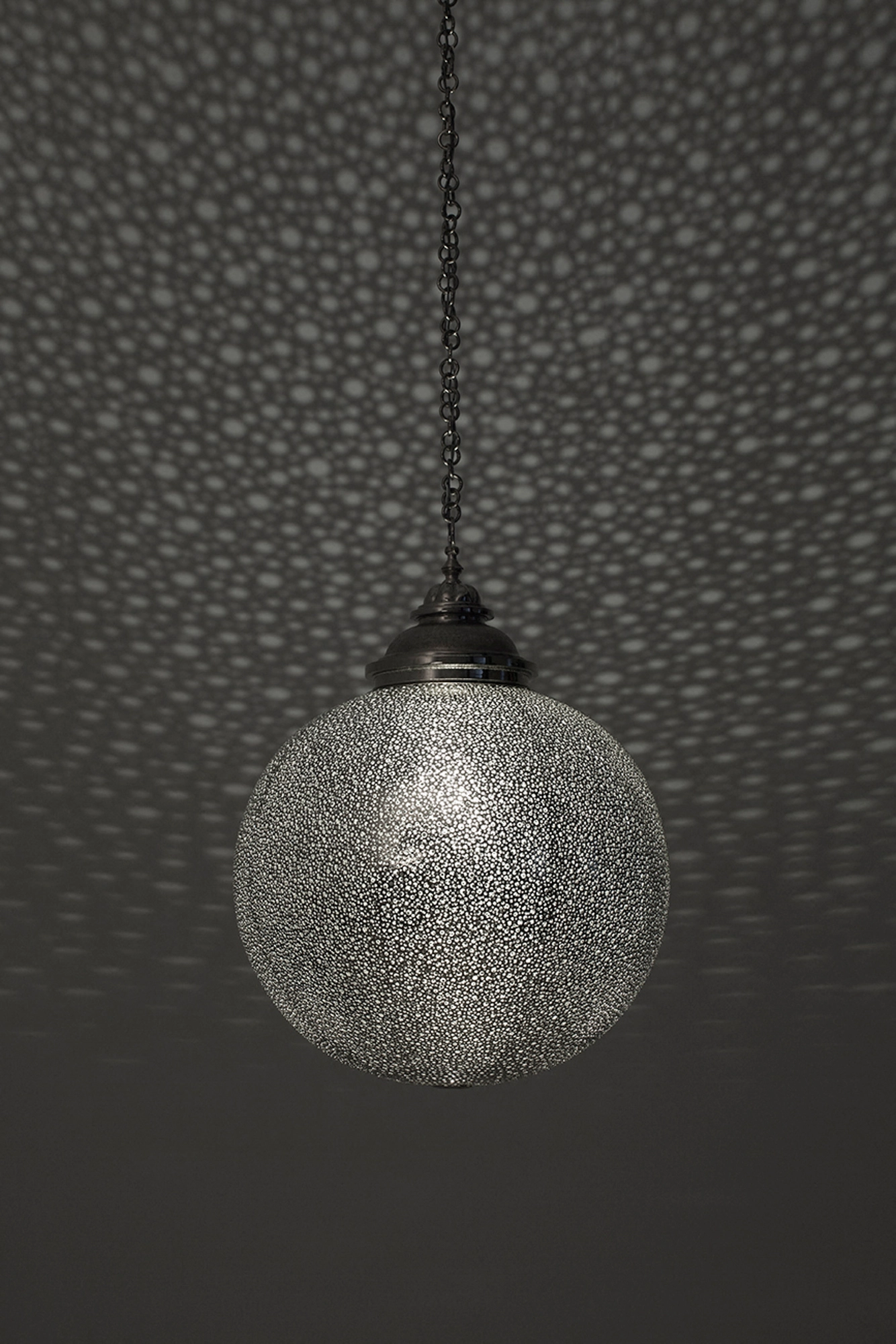 Spherical pierced-silver globe pendant on a chain, glowing against a grey wall flecked with light dots.