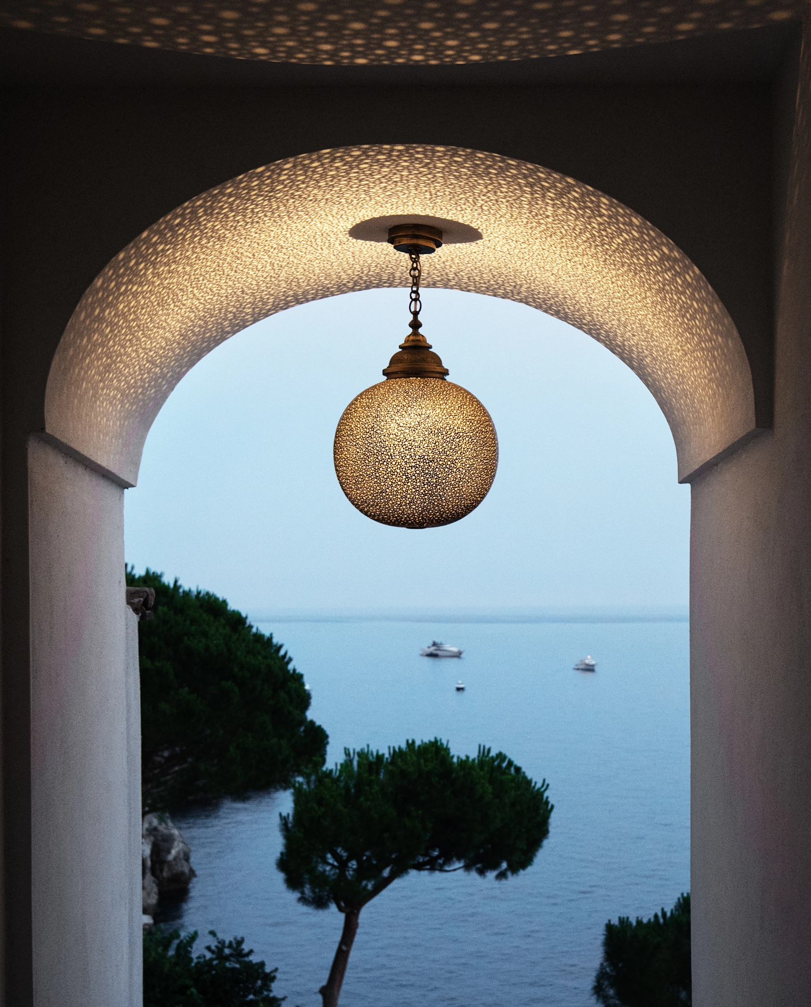 Brass globe pendant beneath an arch framing a sea view with an umbrella pine tree.