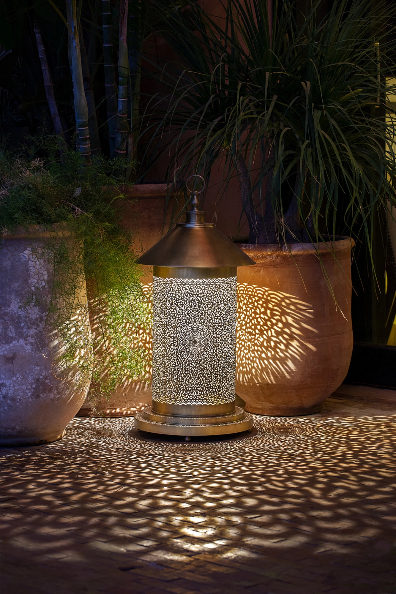 A pierced cylindrical floor lamp glowing among garden planters at night, scattering dotted light on the ground.