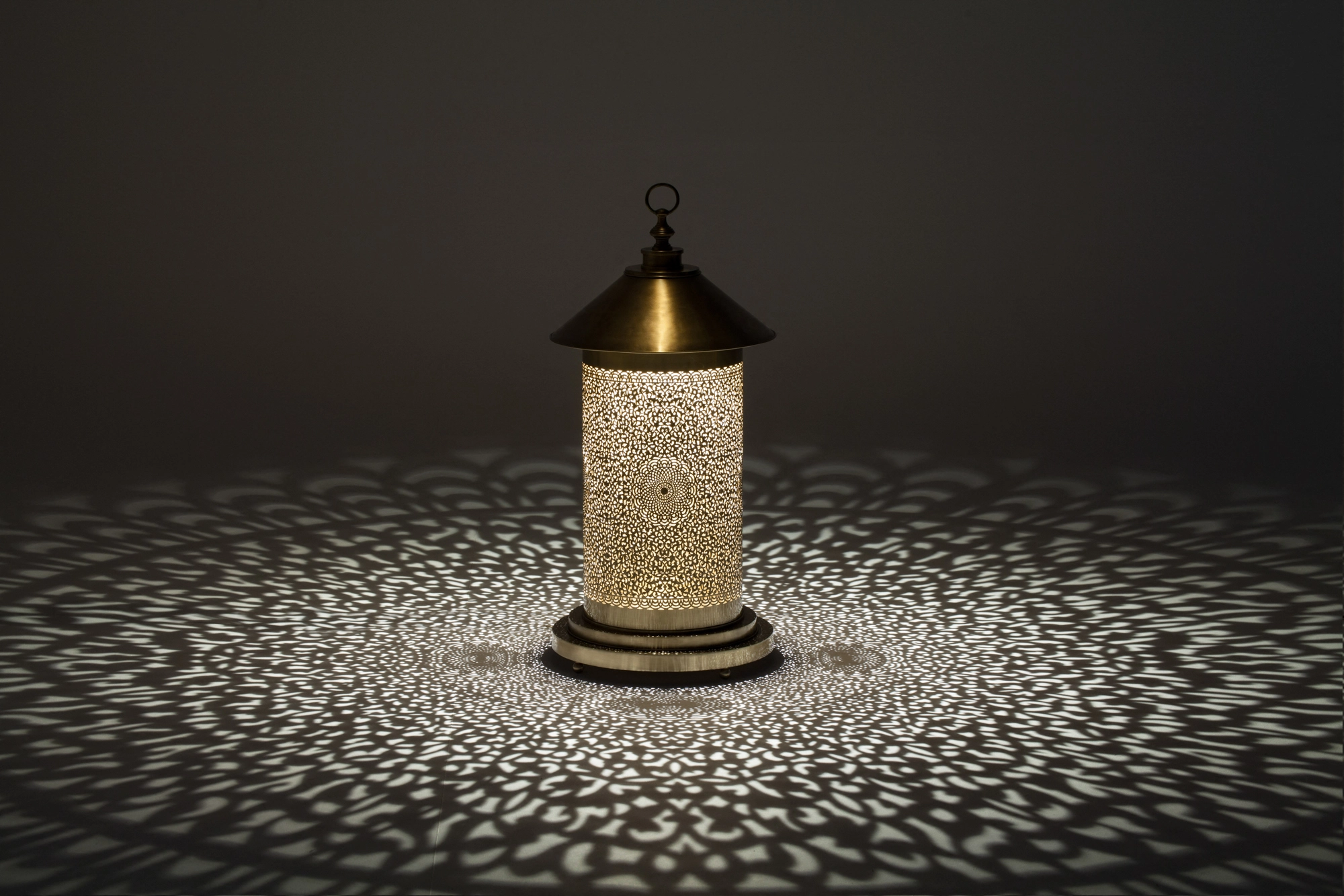 A small pierced cylindrical floor lamp with a pointed top, scattering dotted light across a grey floor.