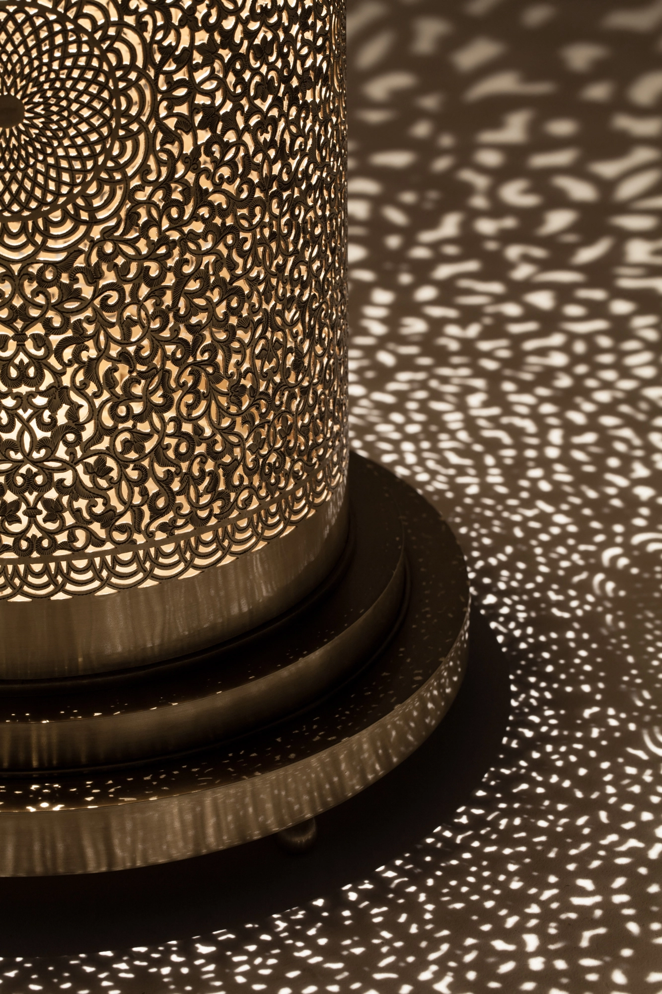 Close-up of a pierced cylindrical floor lamp's lower body and tiered base amid a spray of dotted light.