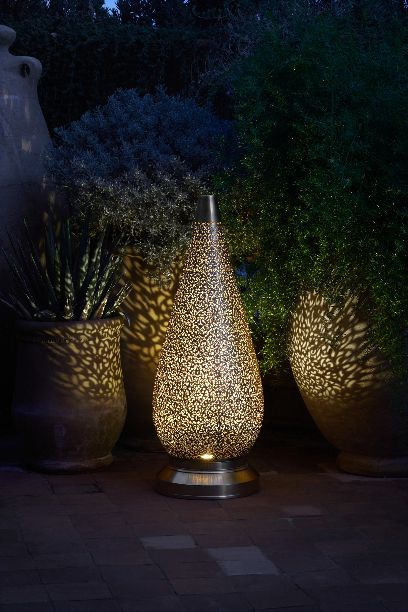 A pierced teardrop floor lamp glowing among garden planters at night, casting light on the foliage.