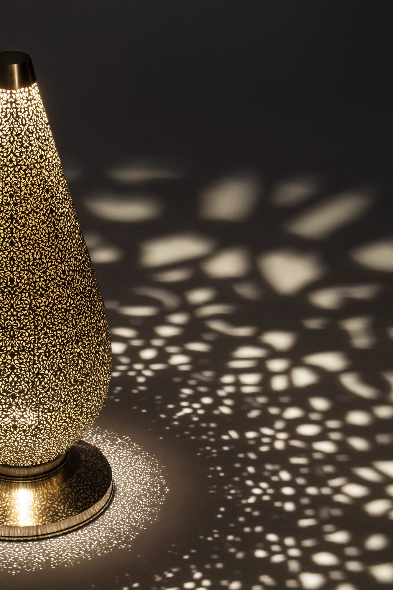 A pierced teardrop floor lamp lit on its base, scattering a warm dotted spray across the floor.