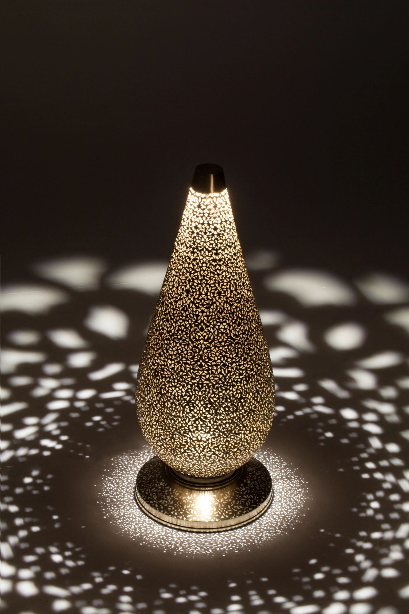 A pierced teardrop floor lamp glowing on its round base within a halo of dotted light.