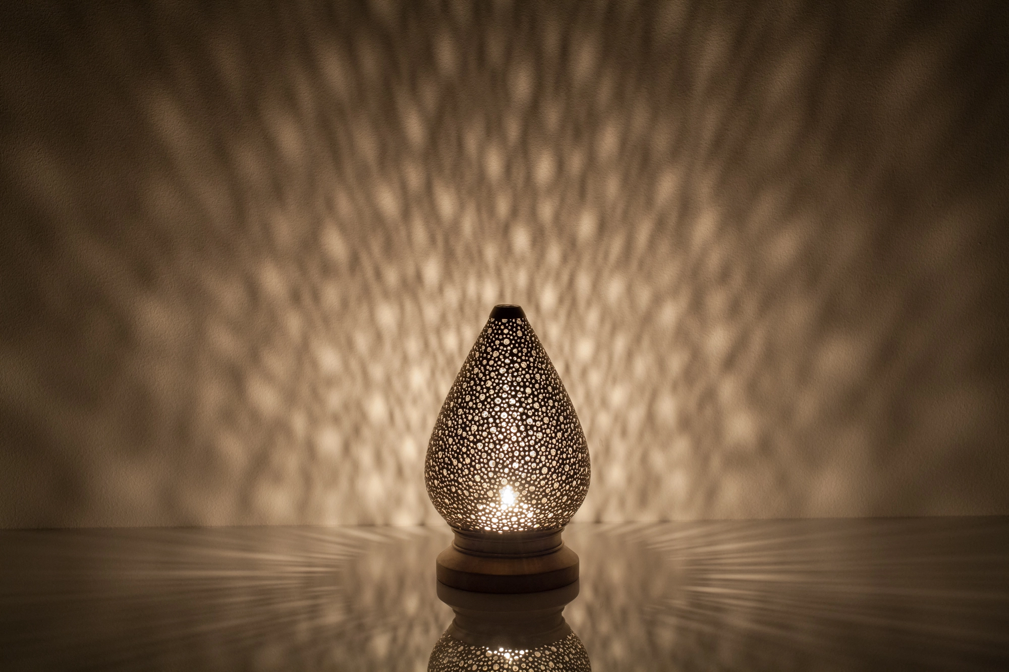 A small teardrop-shaped pierced metal candle holder glowing warmly above its reflection on a dark surface.