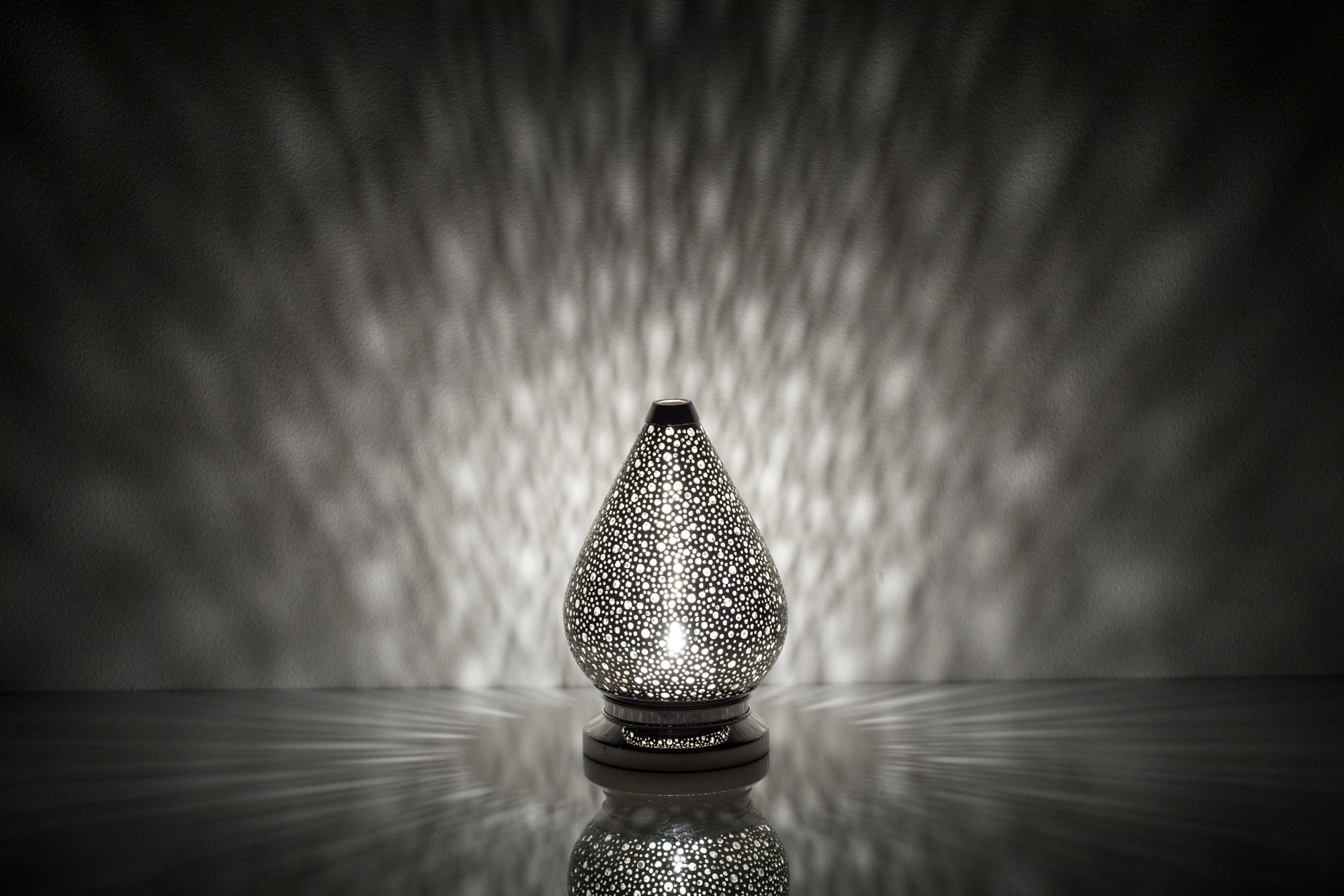 A small teardrop-shaped pierced metal candle holder scattering pale dappled light above its reflection.