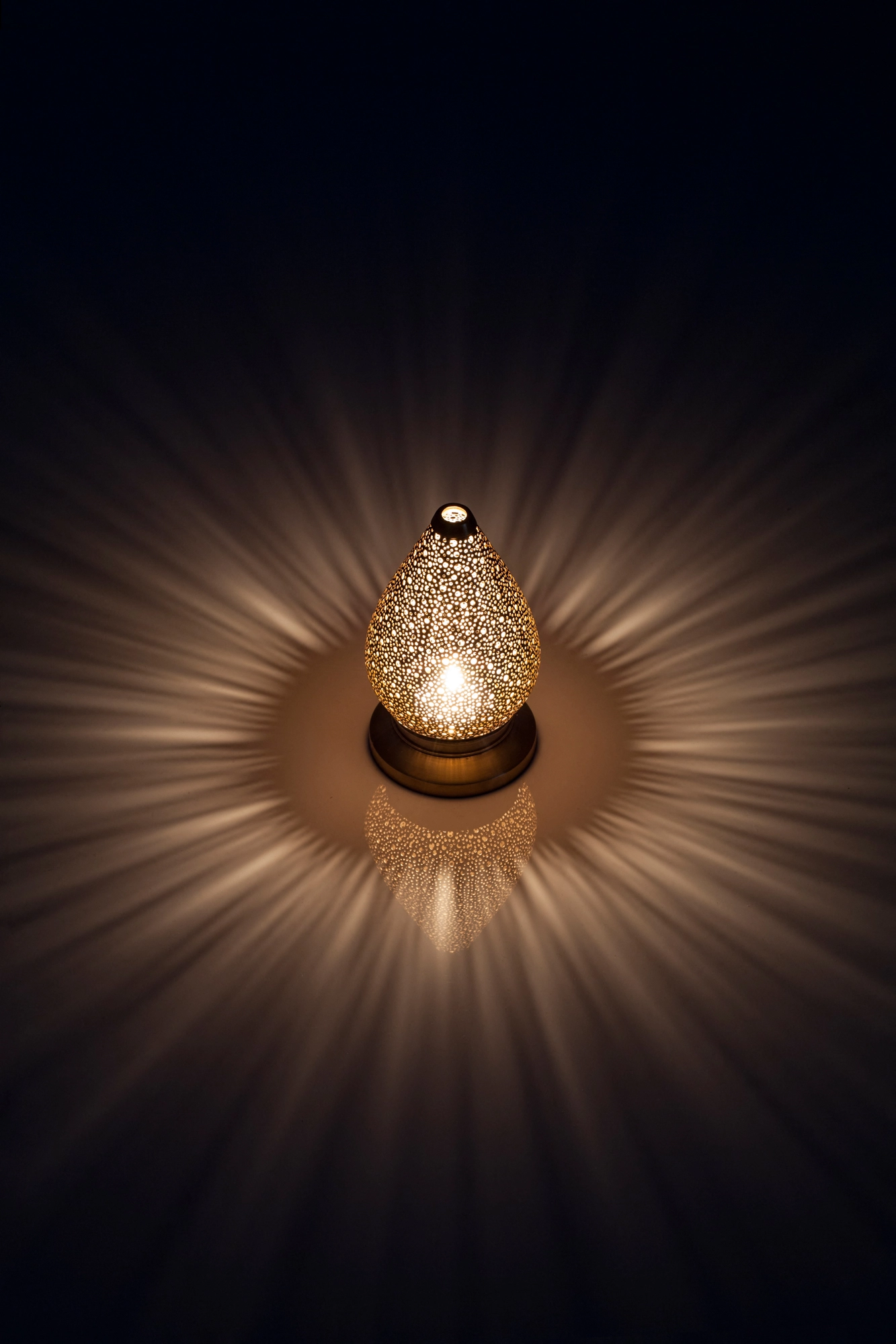 A small teardrop-shaped pierced candle holder casting a warm radial burst of light against a dark background.