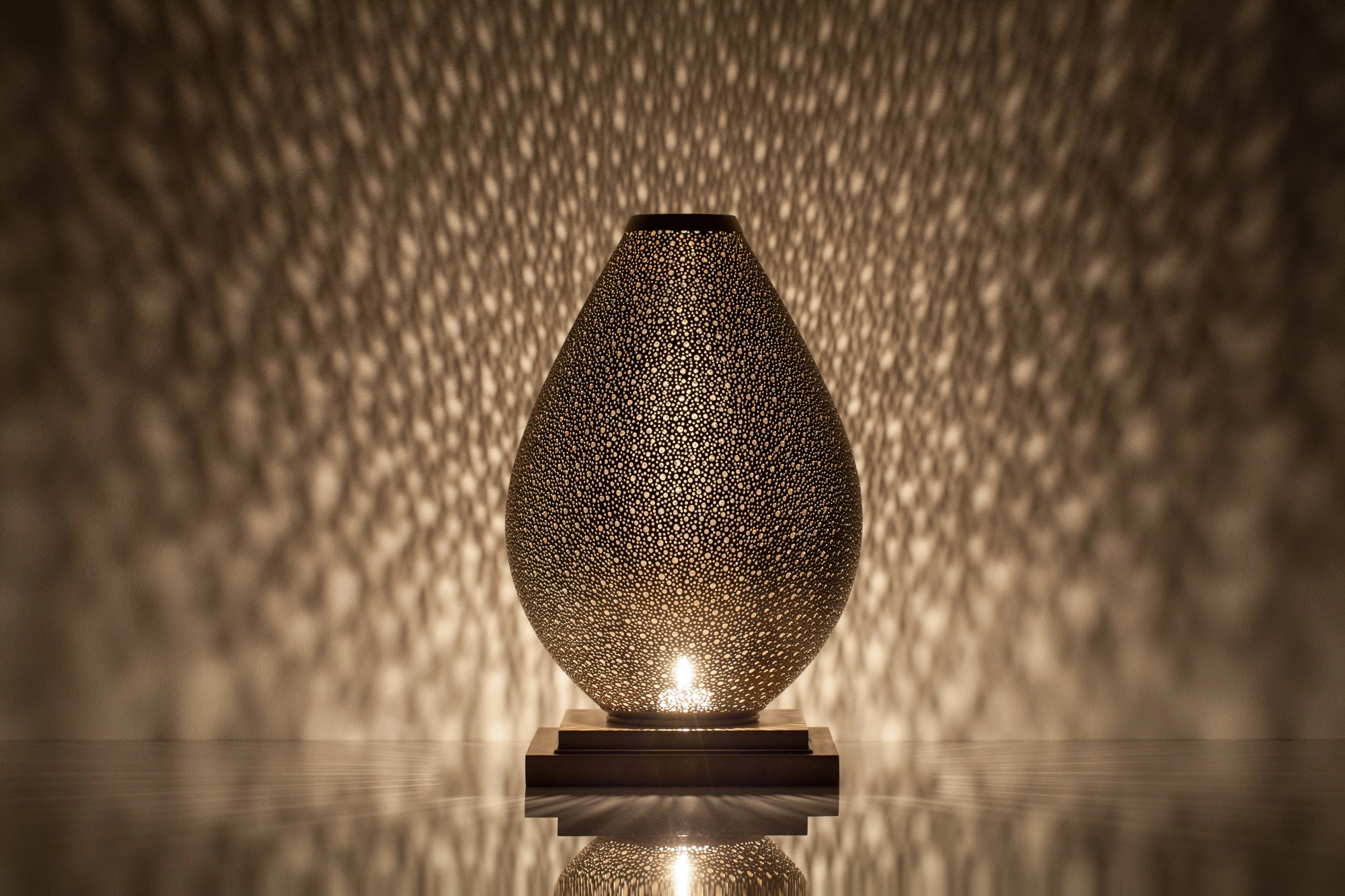 An egg-shaped pierced metal candle holder glowing warmly above its reflection on a dark surface.