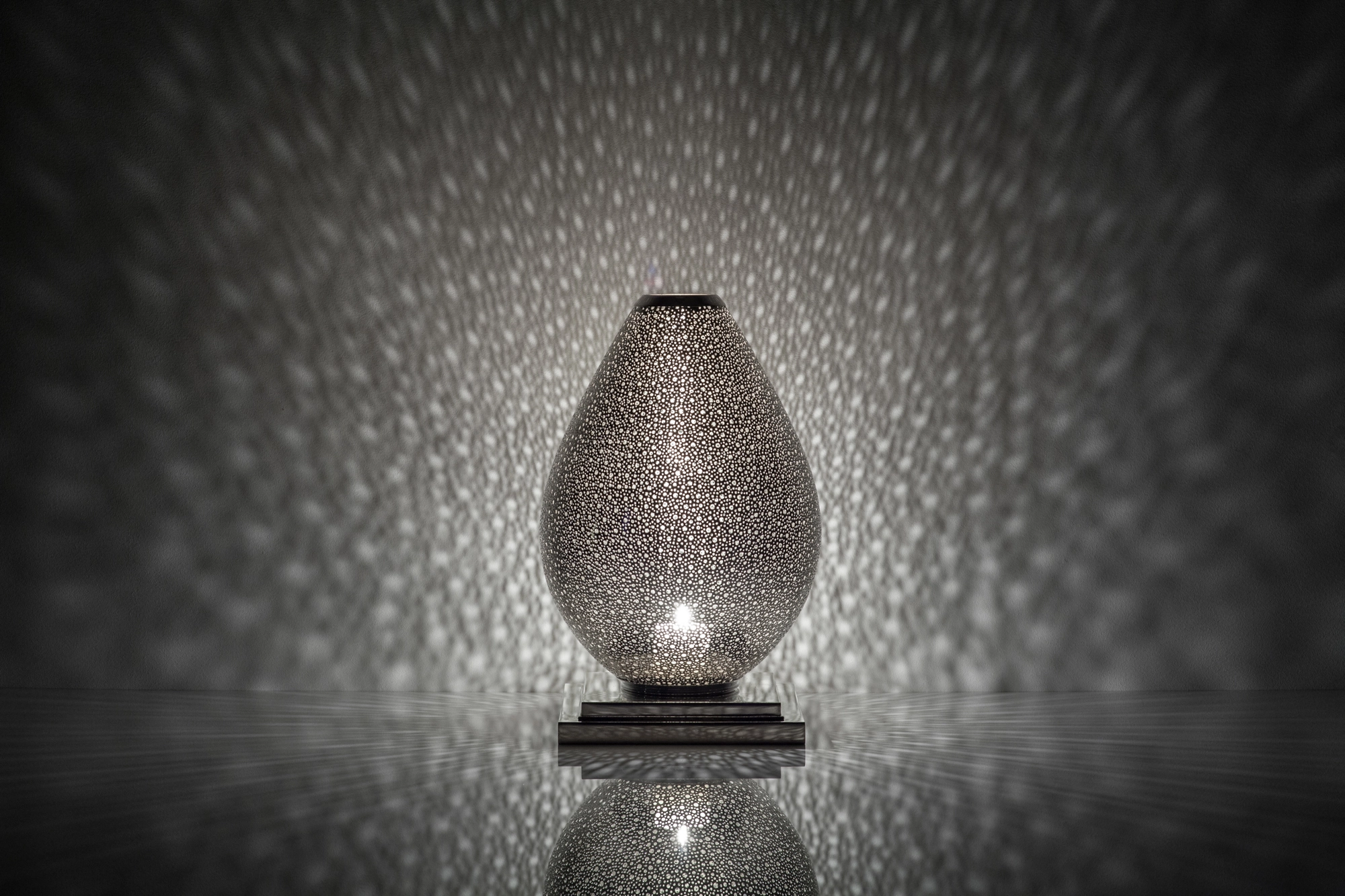 An egg-shaped pierced metal candle holder in silver, scattering soft dappled light above its reflection.