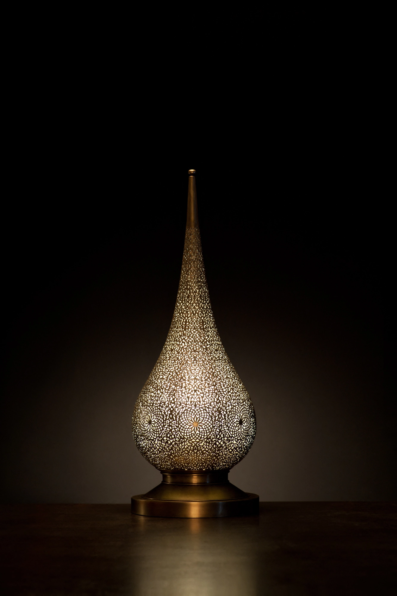 A tall teardrop-shaped pierced metal candle holder glowing softly on a dark surface against a black background.