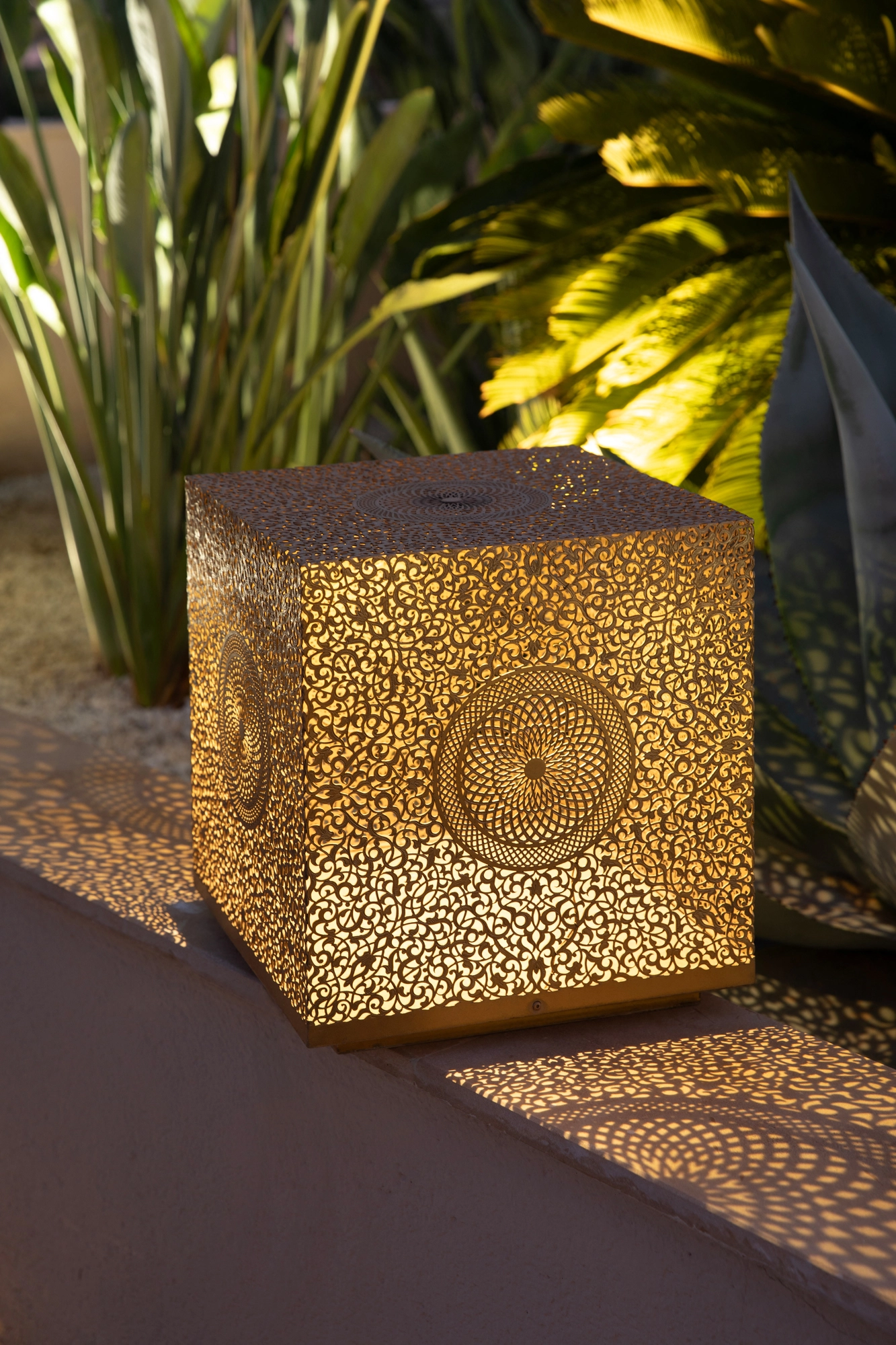 Pierced brass cube floor lamp glowing on the ground beside green foliage at night.