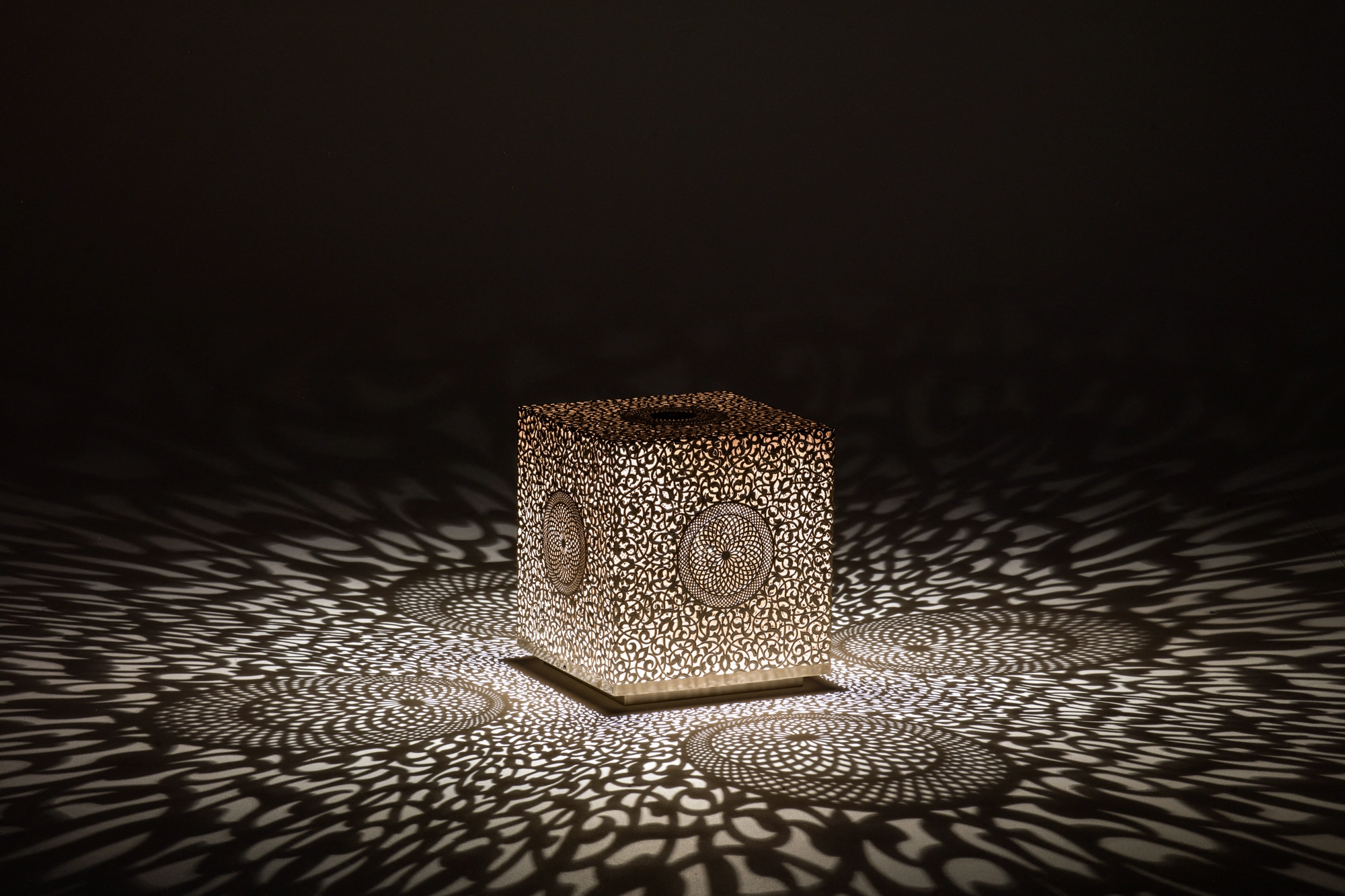 A pierced cube lamp glowing on a dark surface, scattering a fine dotted spray of light around it.