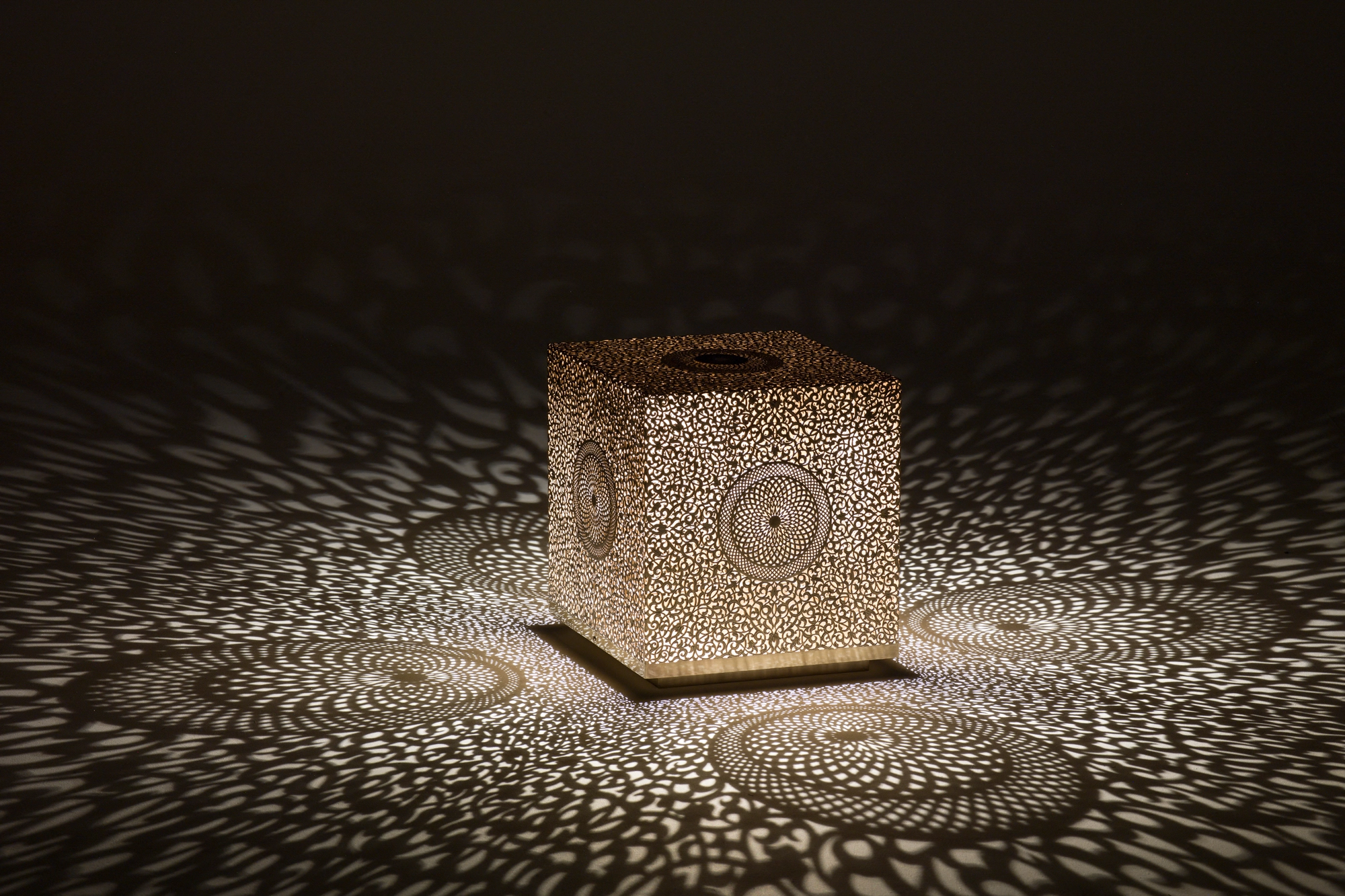 A pierced cube lamp glowing on a dark surface amid a soft spray of dotted light.