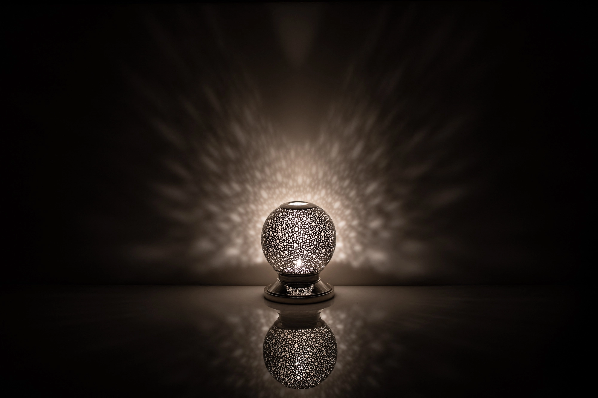 A small spherical pierced metal candle holder glowing dimly above its reflection on a dark surface.