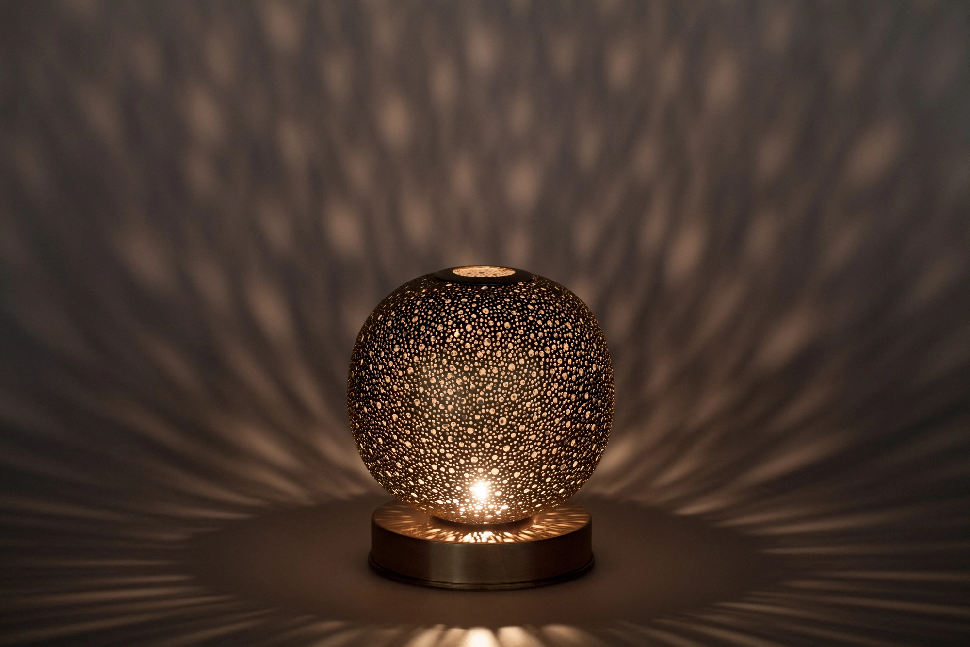 A small spherical pierced candle holder scattering warm dappled light across a dark surface.