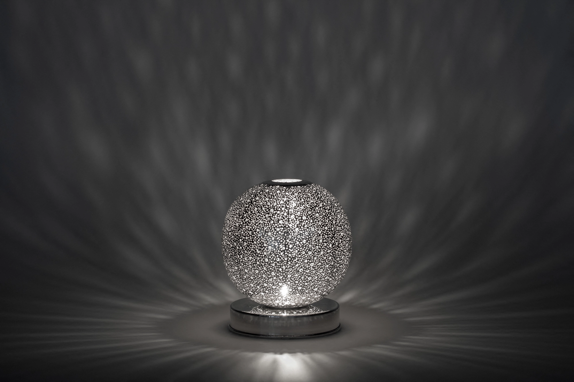 A small spherical pierced candle holder casting soft rippling light above its reflection on a grey surface.