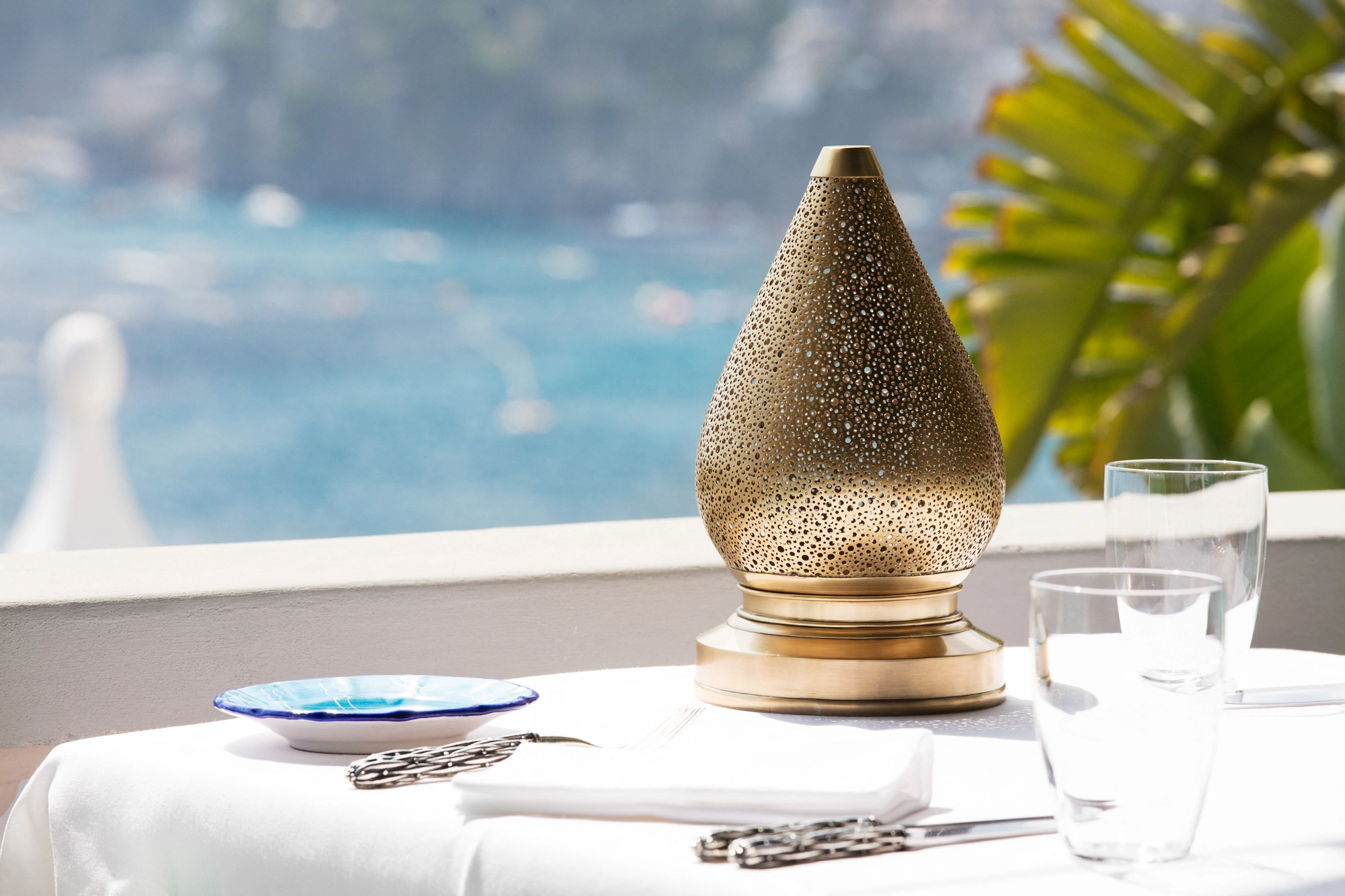 Gold pierced teardrop lamp on a white-clothed table set beside a pool and open sea view.
