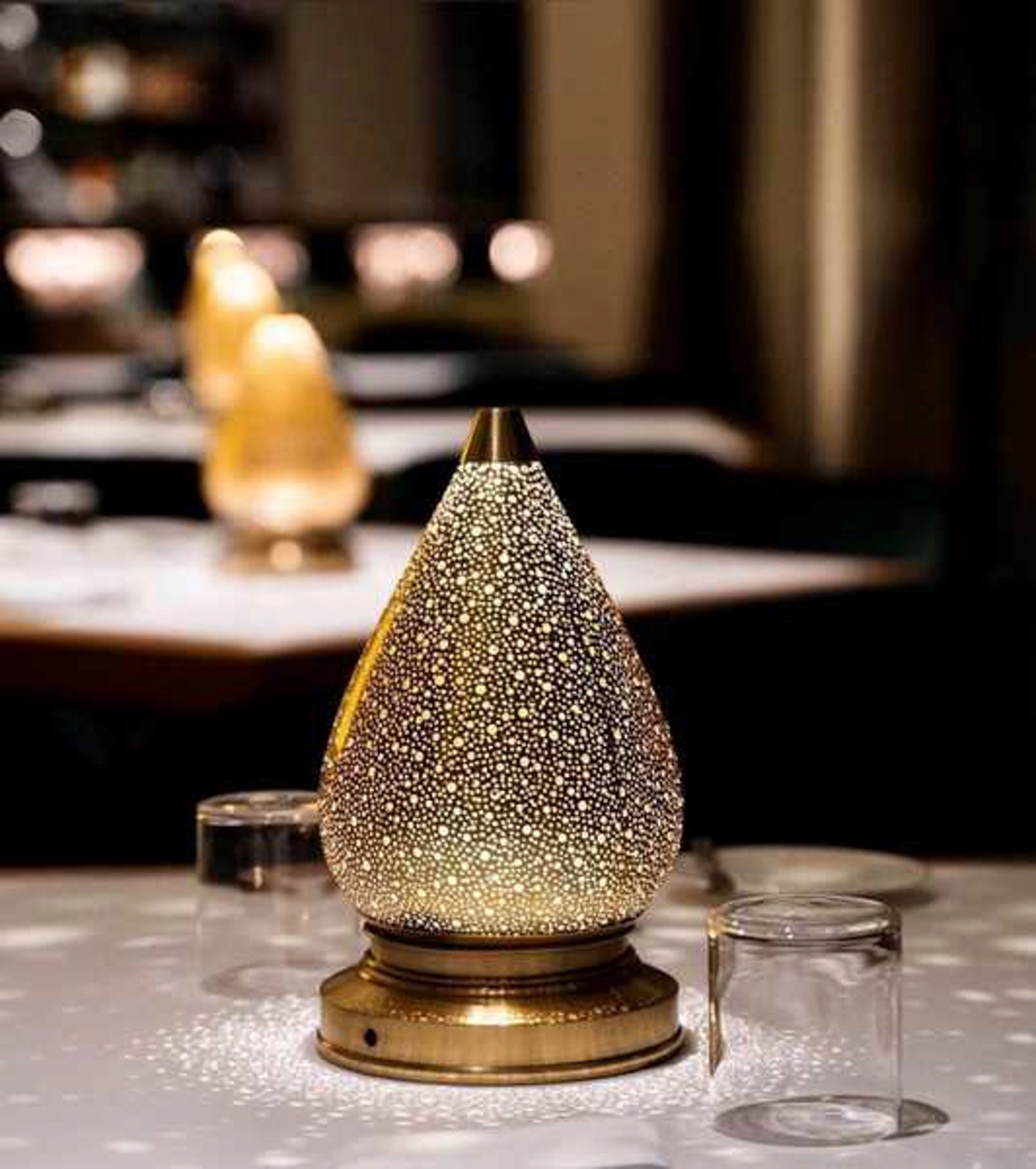 Teardrop pierced lotus table lamp glowing on a set restaurant table at night, among glassware.