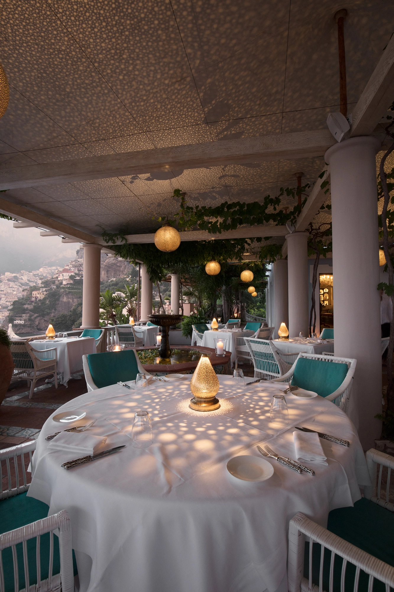 Dusk restaurant terrace with a glowing teardrop lamp on a round table scattering light patterns.
