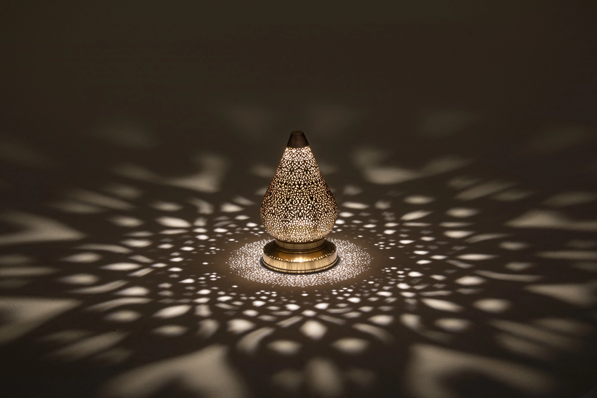 Small pierced lotus lamp glowing in a dark room, casting a wide circular halo of light dots.