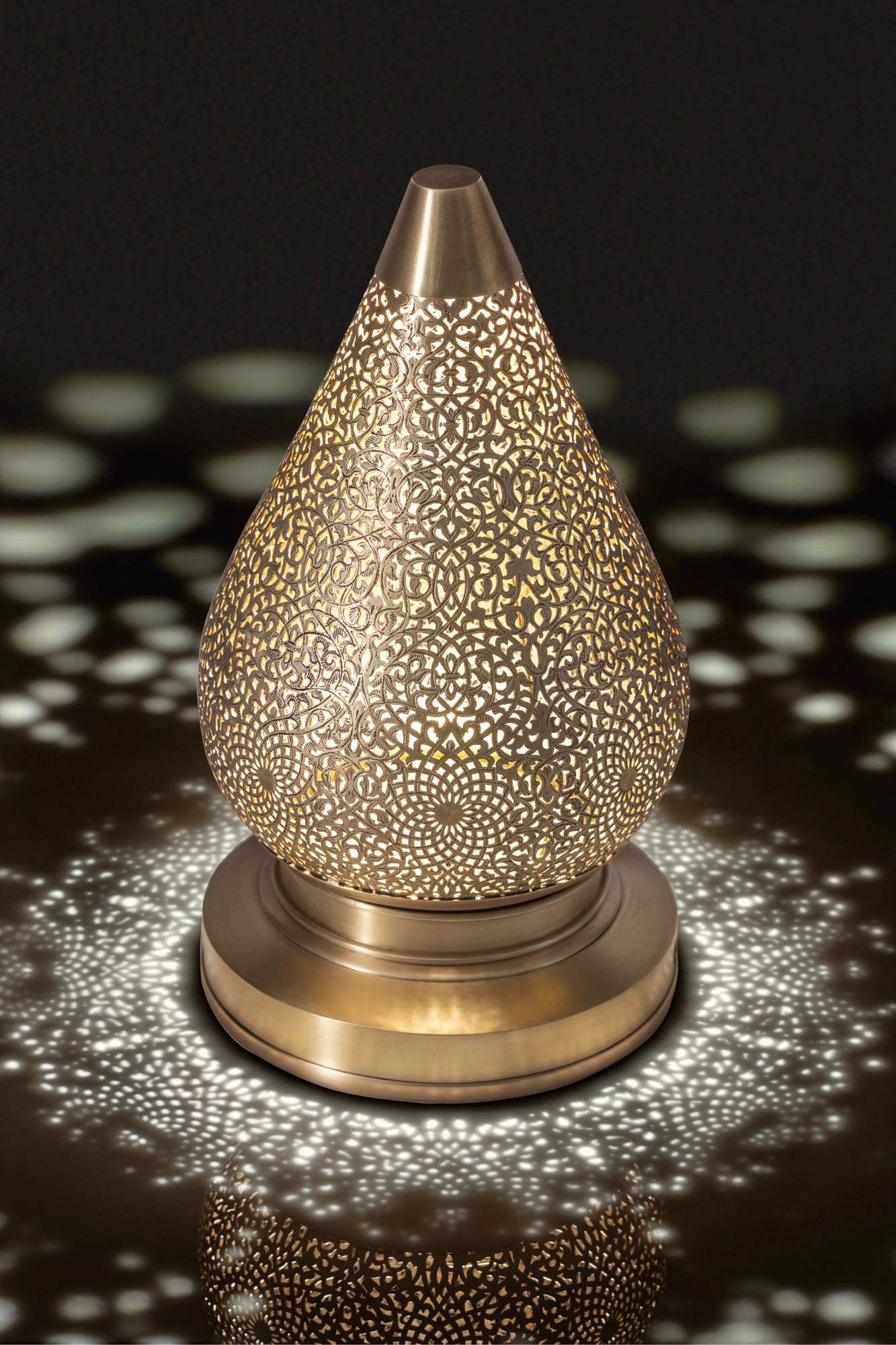 Teardrop-shaped pierced-brass lotus lamp on a round base, glowing and casting a ring of light dots.