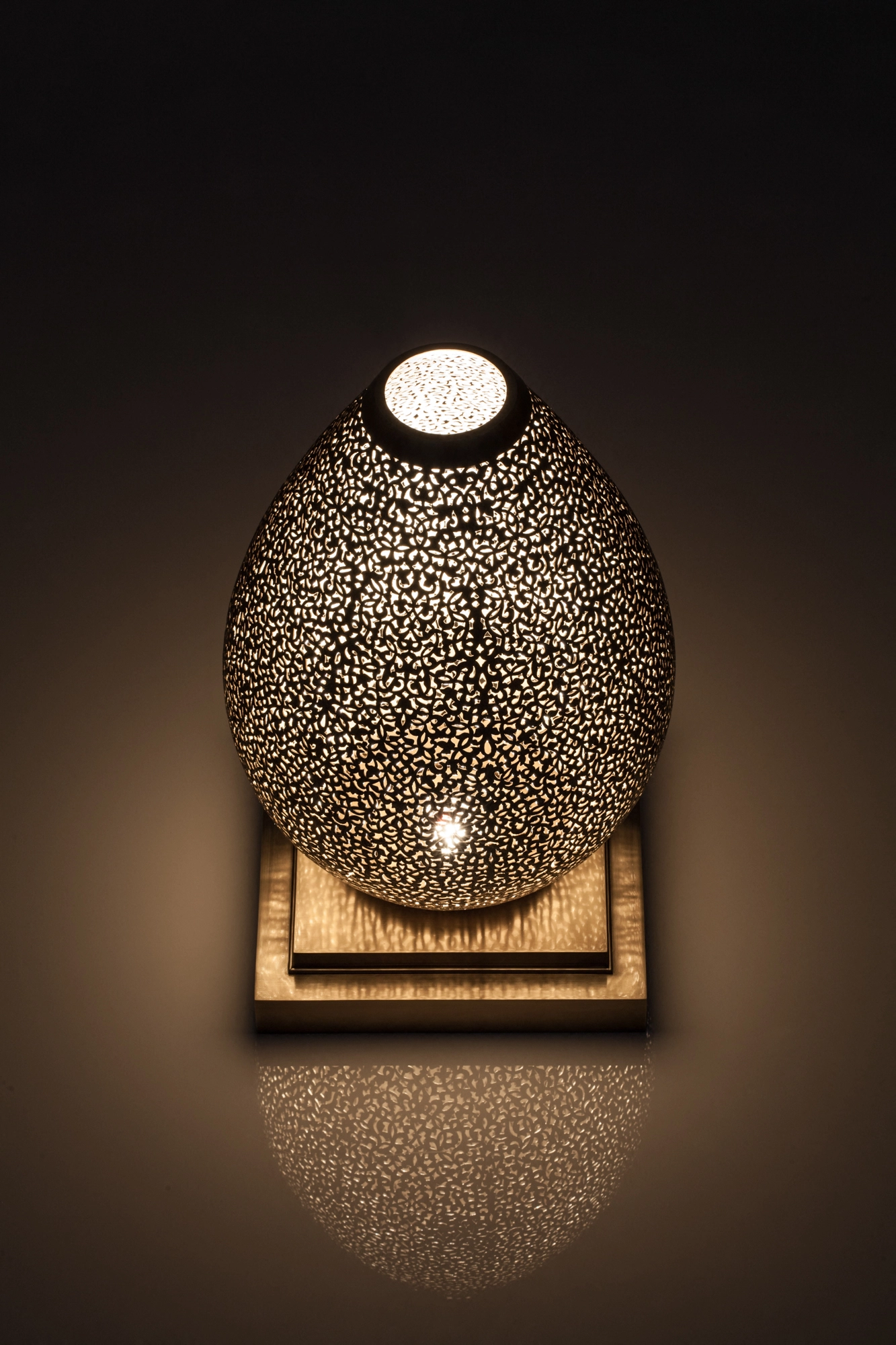 An egg-shaped pierced metal candle holder on a wooden base, glowing warmly with a bright opening at the top.