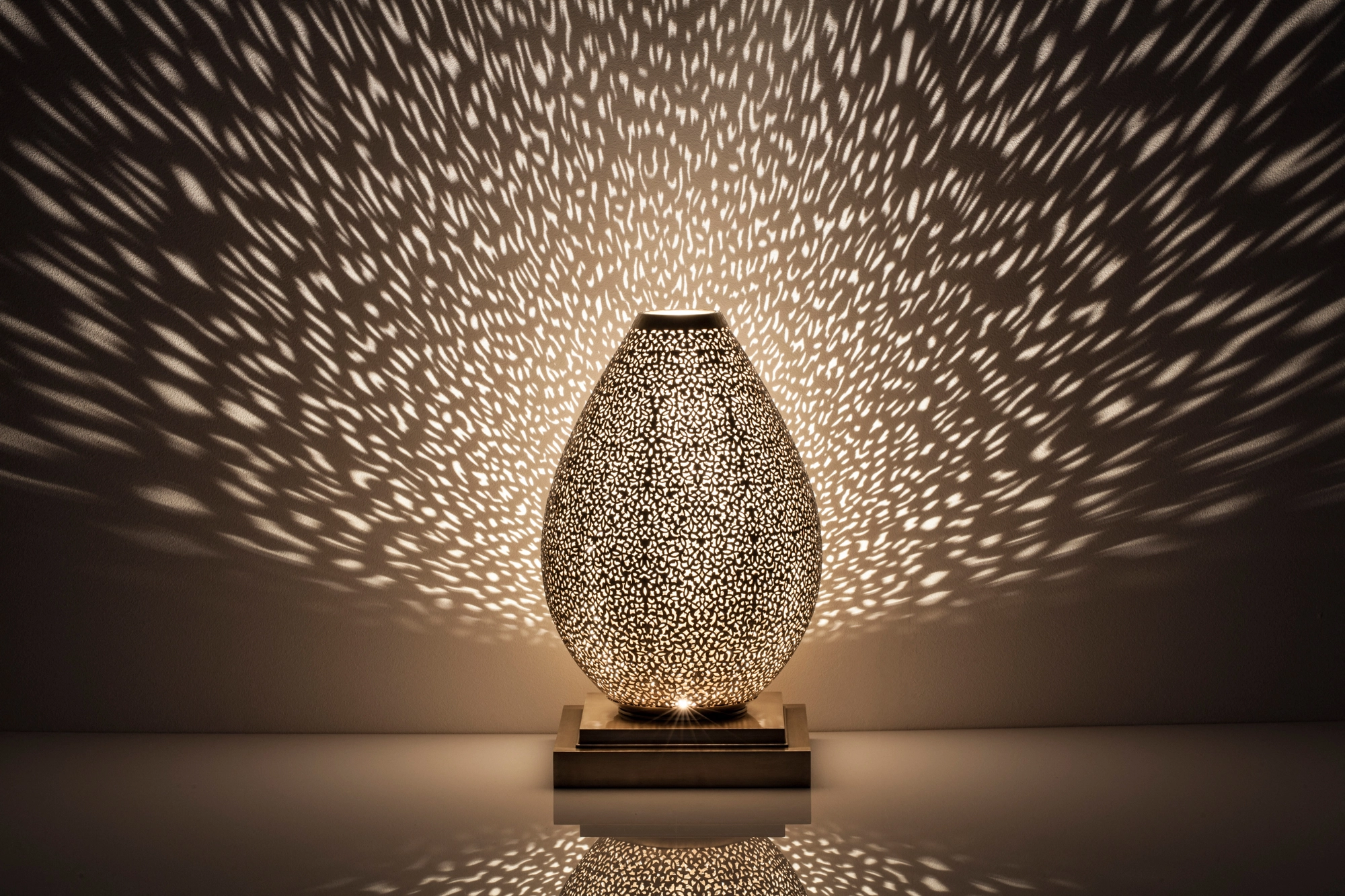 An egg-shaped pierced metal candle holder casting a wide spray of warm dappled light against a dark background.