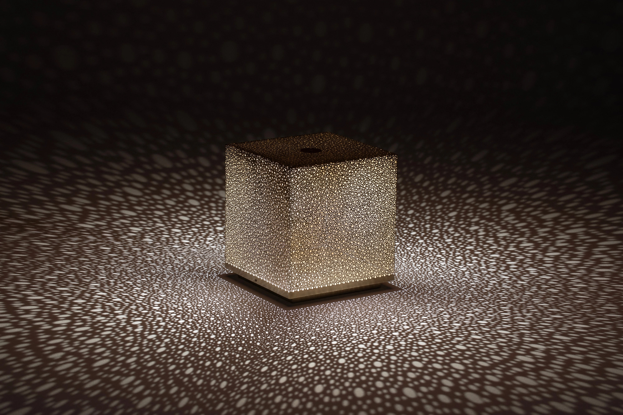 A pierced cube lamp glowing on a dark reflective surface, scattering dotted light around it.