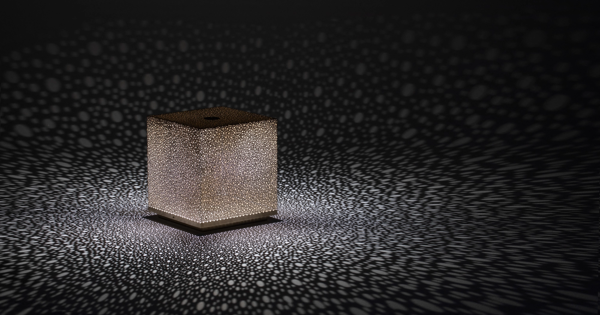 A small pierced cube lamp glowing on a dark surface within a halo of dotted light.