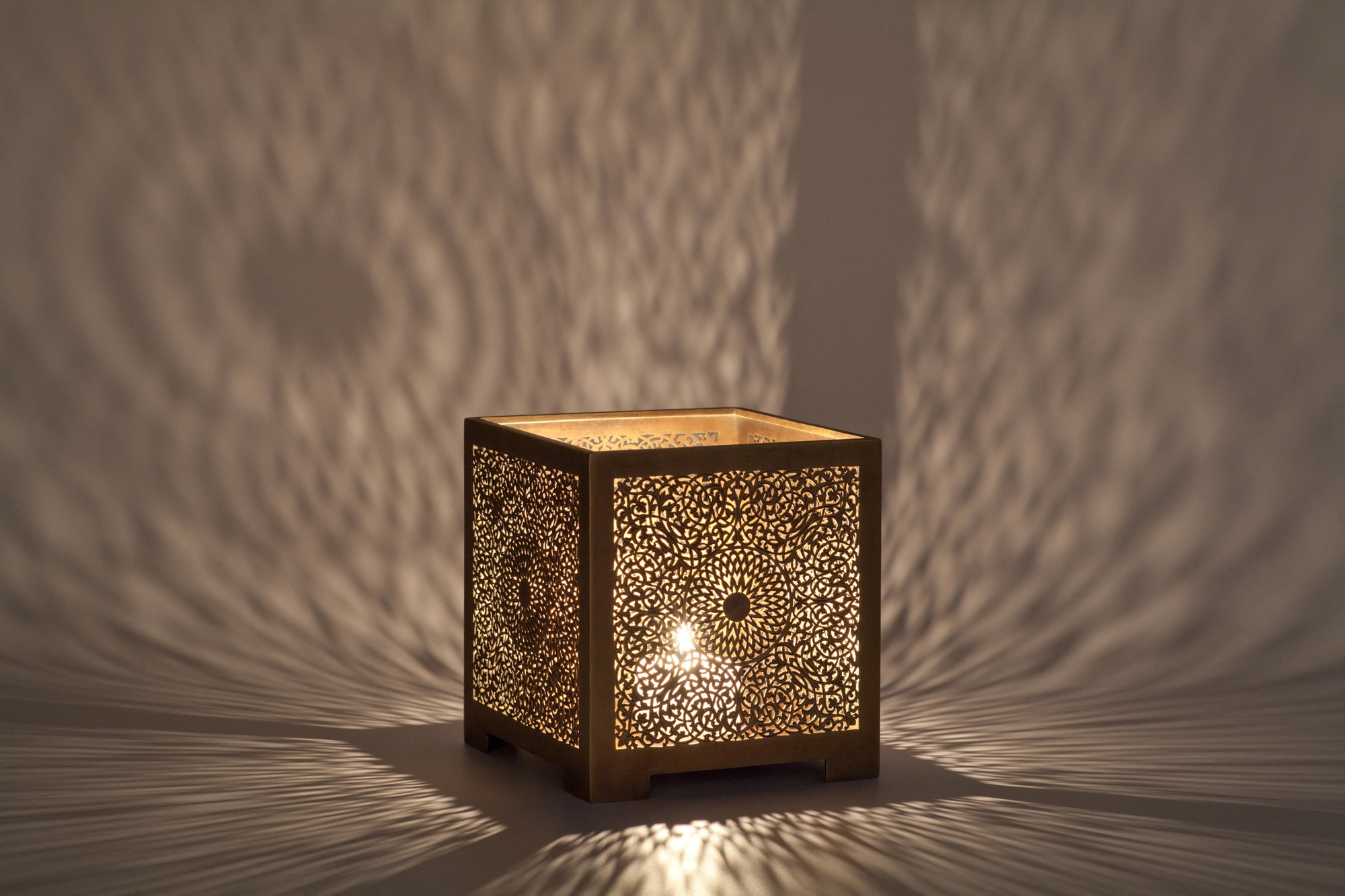 A square pierced metal candle box lit from within, casting a warm burst of dappled light onto the surface around it.