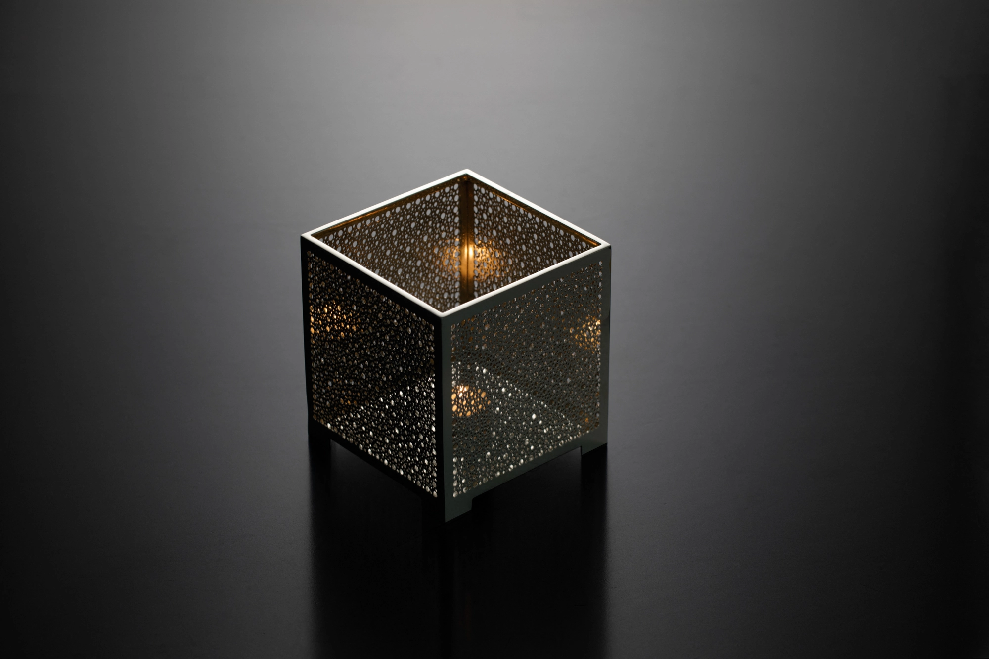 A square pierced metal candle box holding a lit candle, glowing warmly against a dark background.