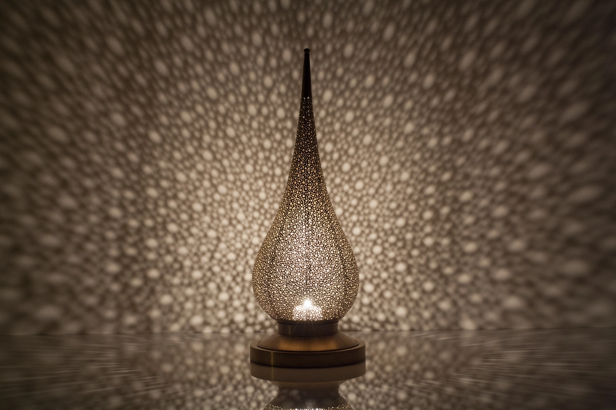 A flame-shaped pierced candle holder casting soft dappled light in warm tones against a pale background.