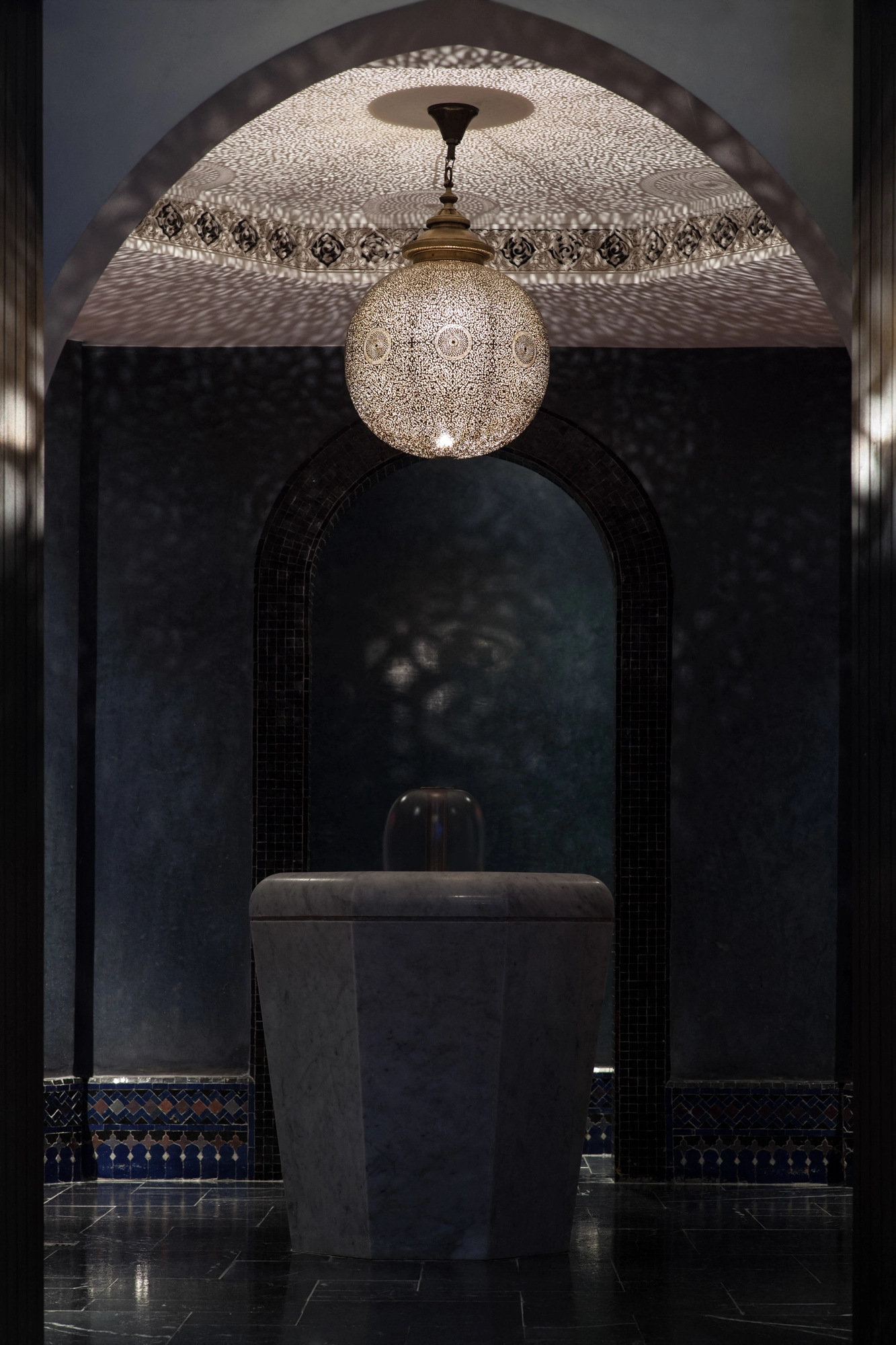 Pierced brass globe pendant glowing in a dark arched alcove, casting dramatic shadows.