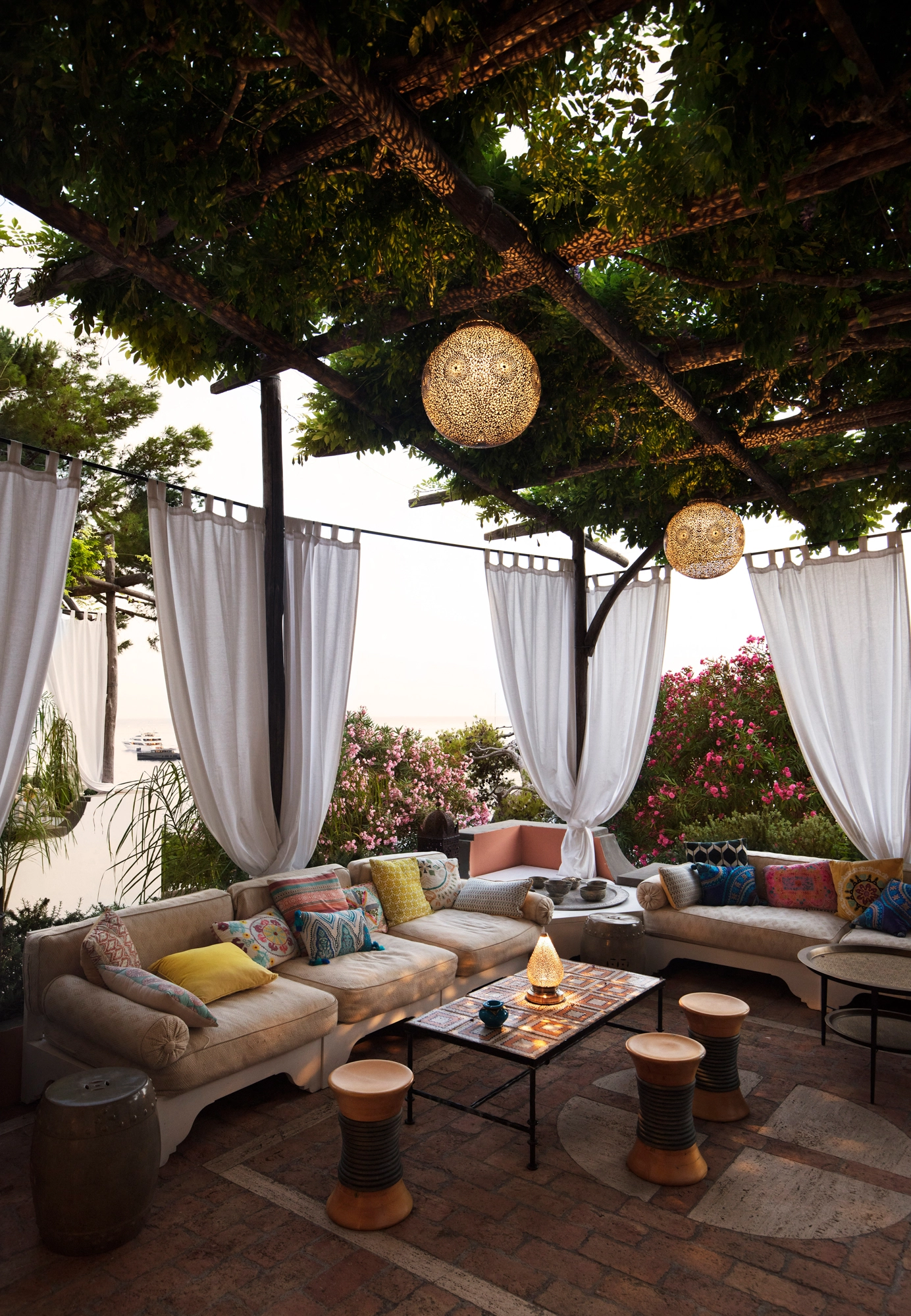 Outdoor terrace lounge with sofas under a pergola, a globe lantern, white drapes and a sea view.