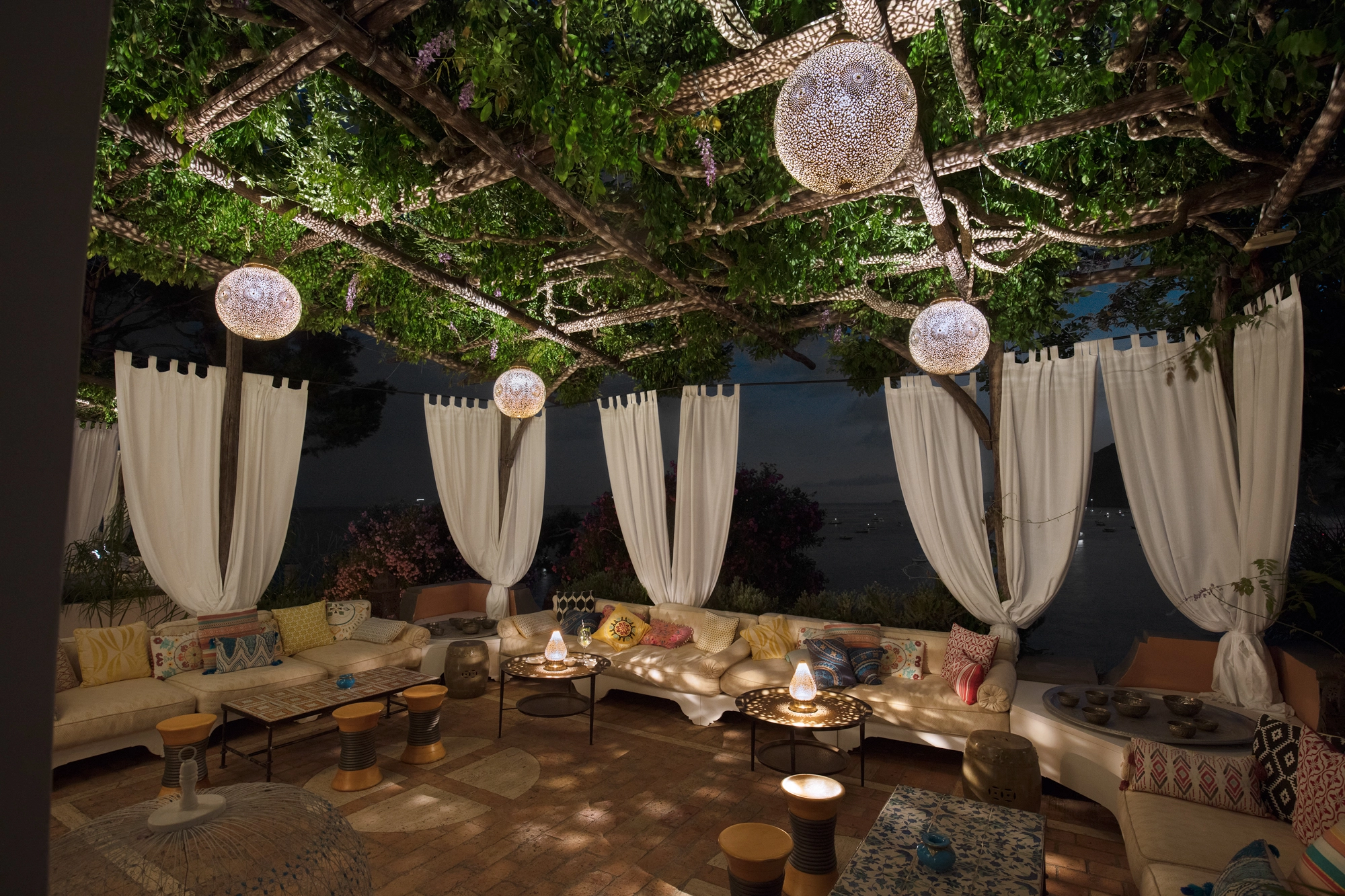 Evening terrace beneath a tree strung with globe lanterns, set for dining above the coast.