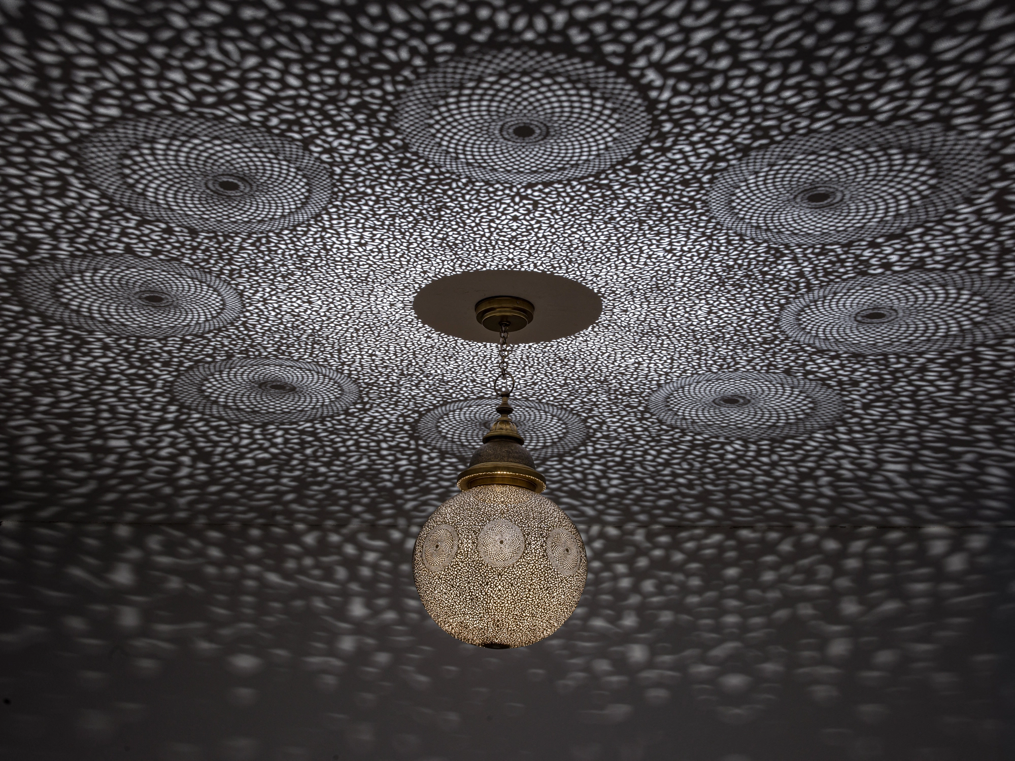 Pierced-brass globe pendant glowing in a dark room, ringed by a wide pattern of disc-shaped light projections.