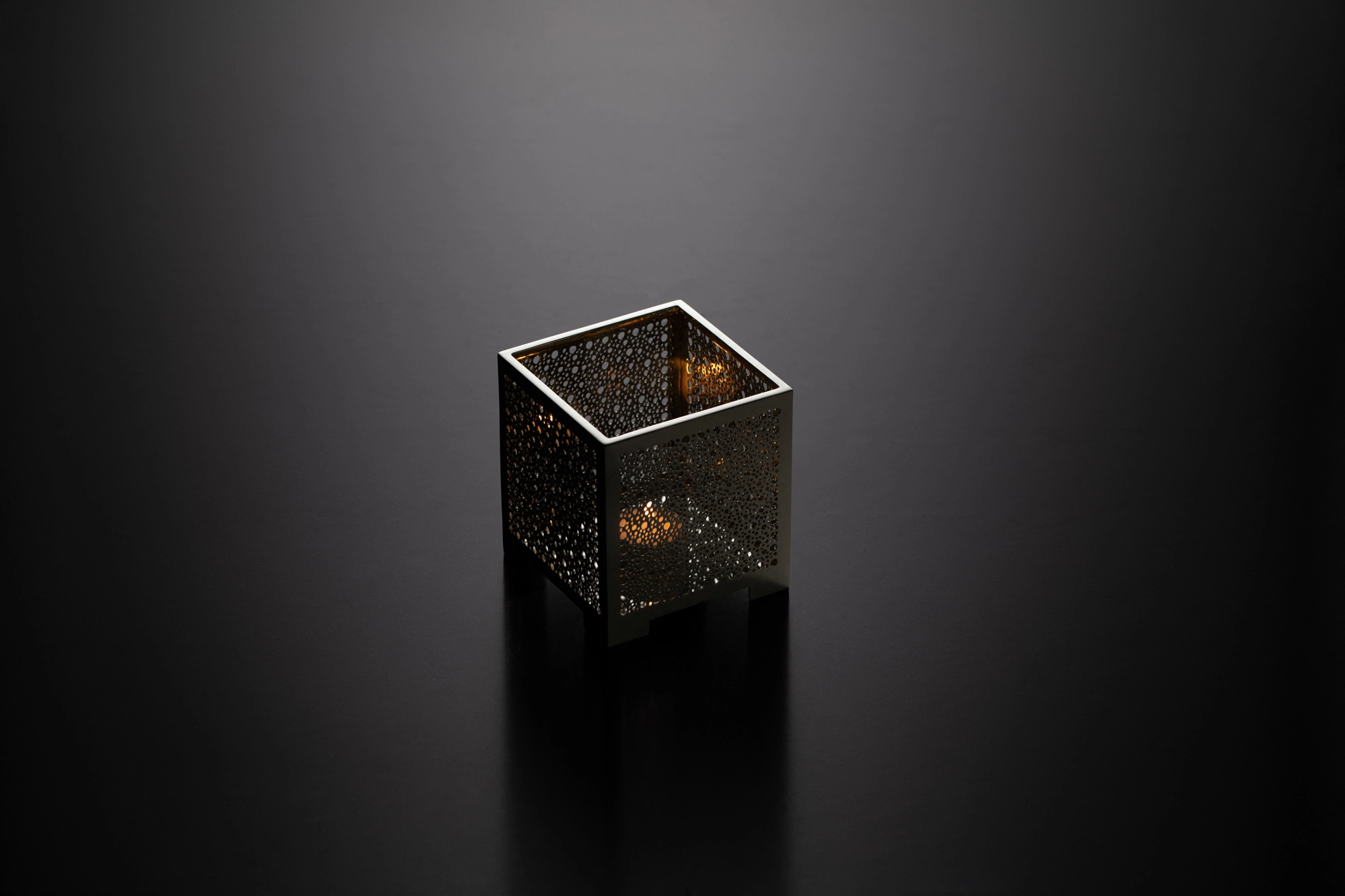 A square pierced metal candle box with a lit candle inside, glowing softly above its reflection on a dark surface.