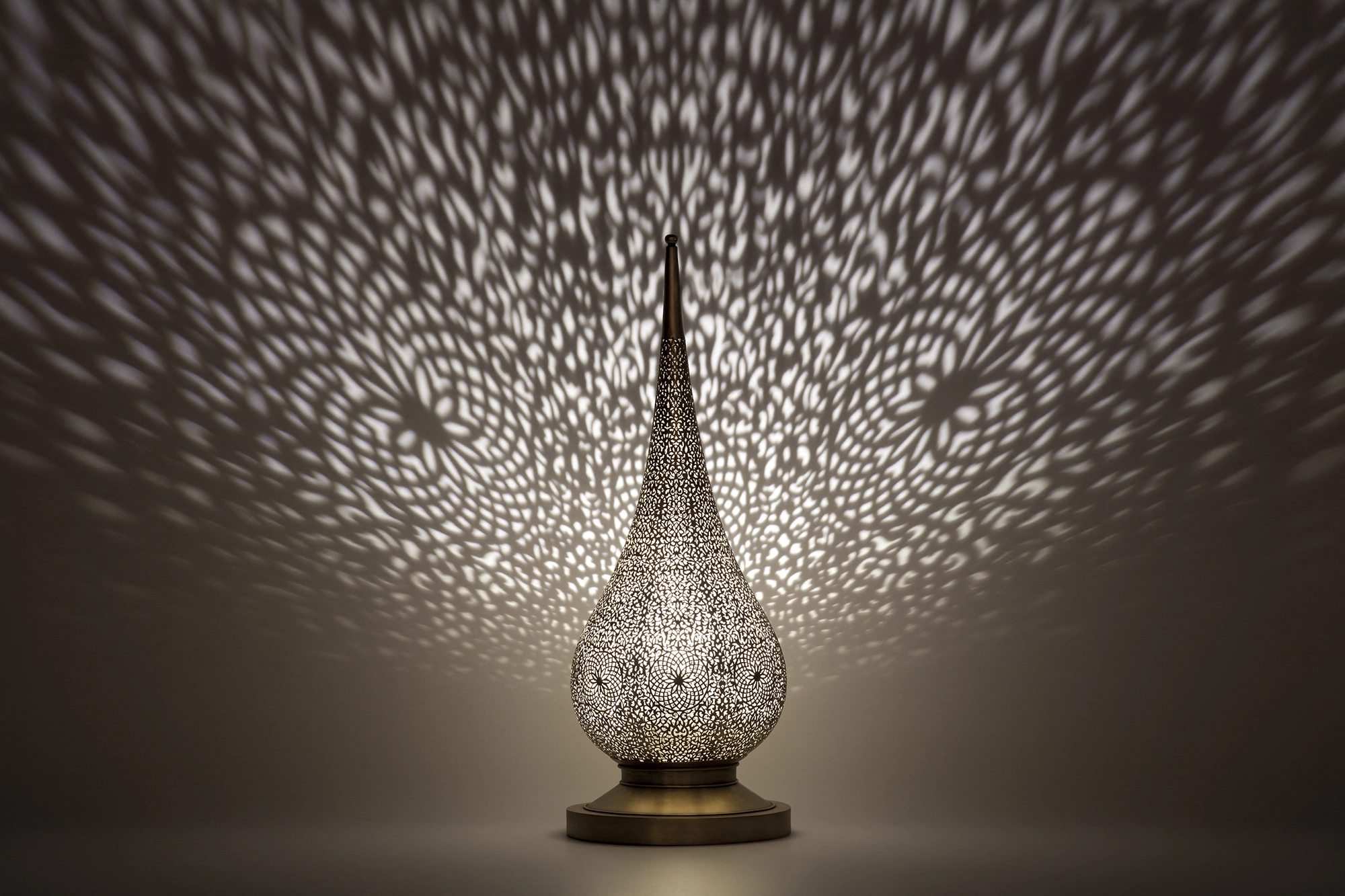 A flame-shaped pierced metal candle holder glowing warmly and scattering dappled light onto the wall behind it.