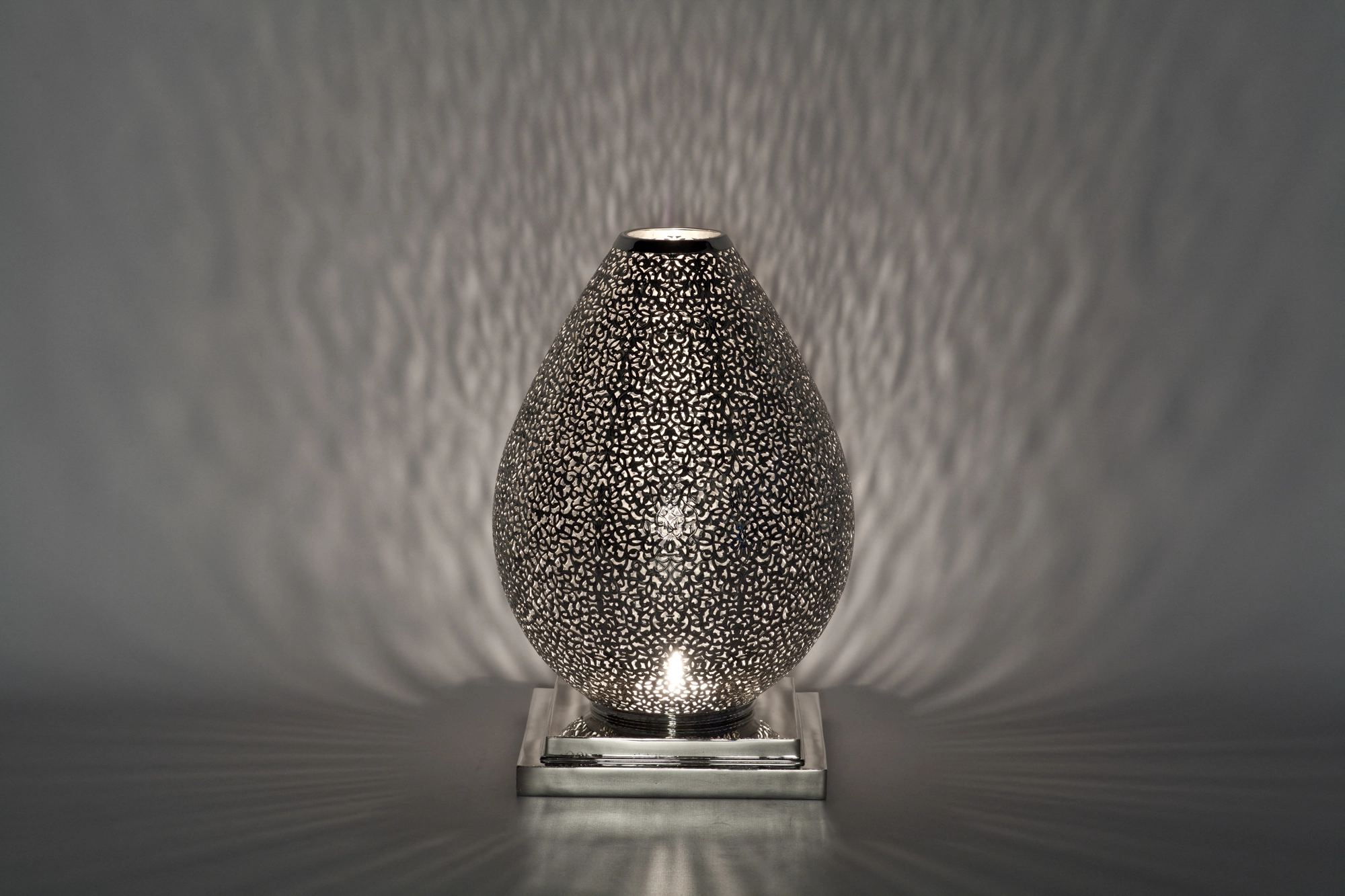An egg-shaped pierced metal candle holder scattering soft dappled light against a pale grey background.