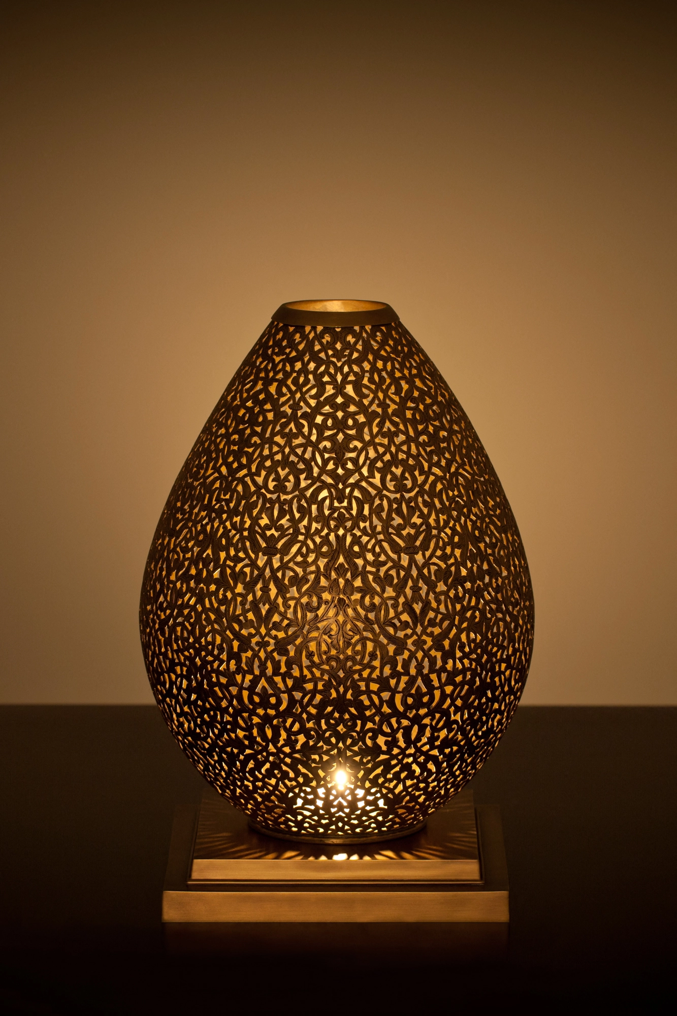 An egg-shaped pierced metal candle holder glowing amber on a wooden base against a dark background.