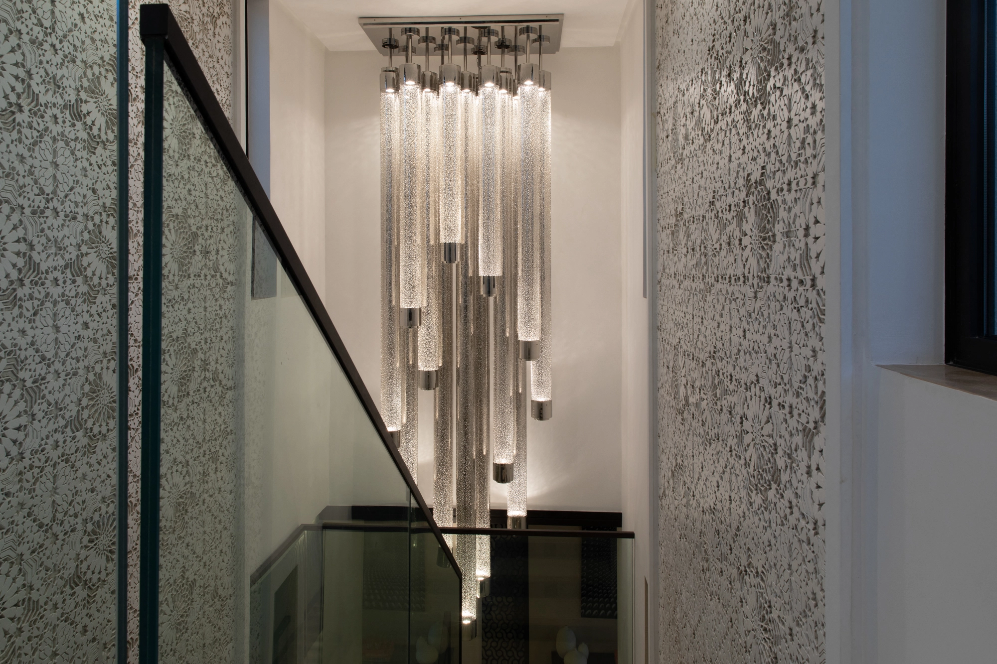 Cylindrical liquid-light glass tubes hanging in a stairwell beside a glass balustrade and stone wall.