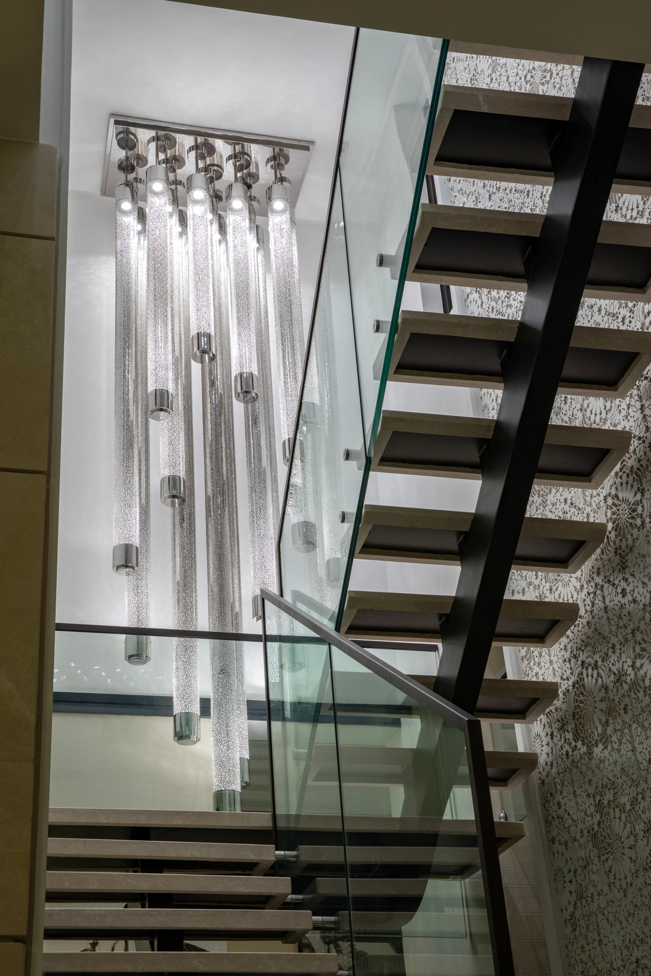 Tall cluster of cylindrical liquid-light glass tubes descending beside a glass-railed staircase.