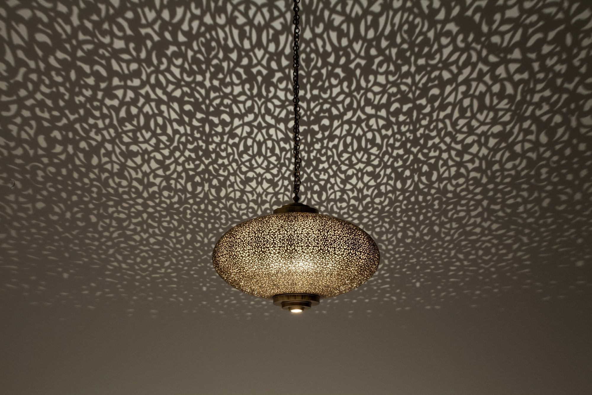 Small pierced-brass squashed-globe pendant glowing on a cord in a dimly lit room.