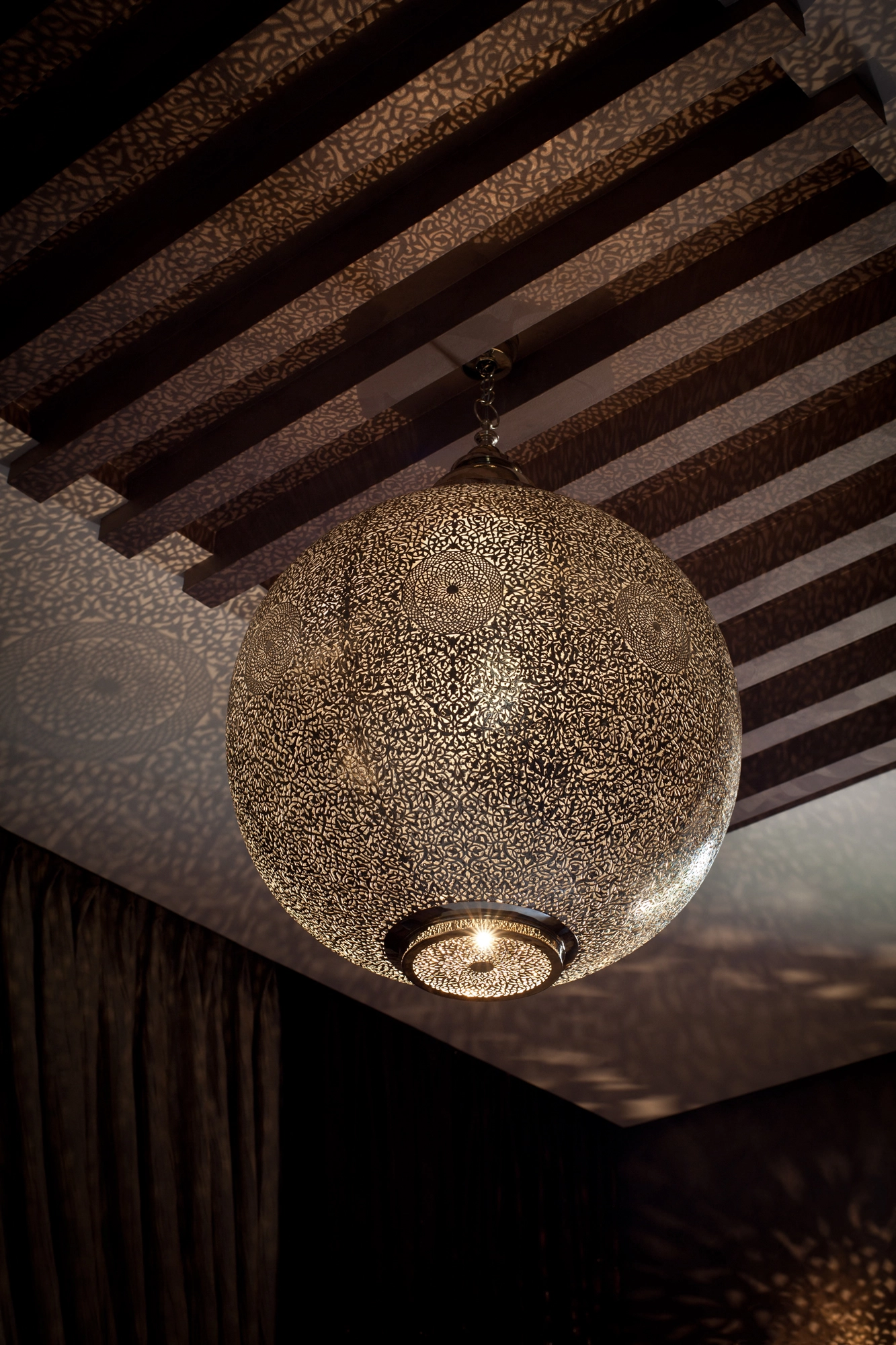 Large pierced-brass globe pendant hanging beneath a dark wood-beam ceiling, glowing and scattering light dots.