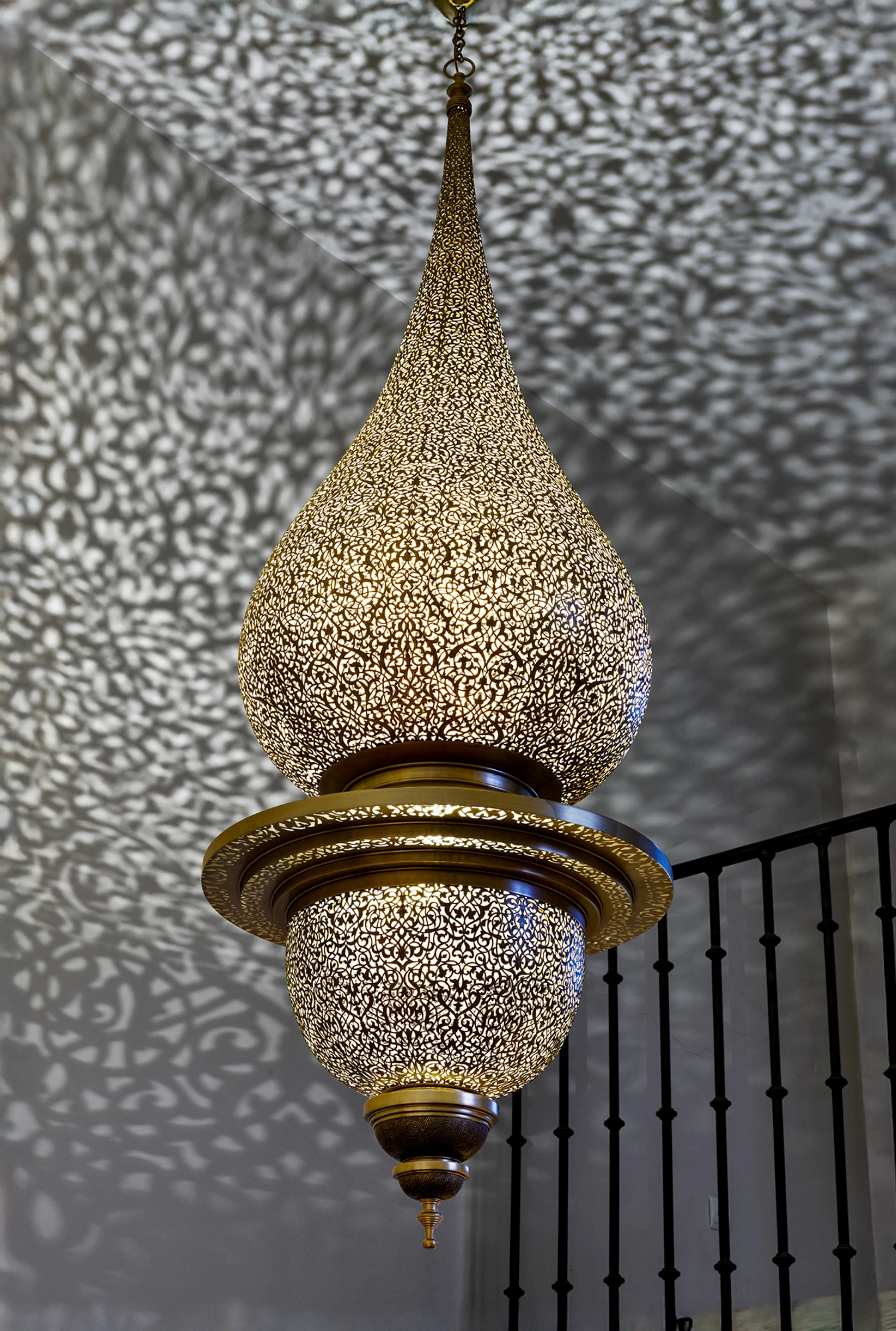 Pierced brass onion-shaped pendant lantern casting lace-like shadows on a wall beside an iron railing.